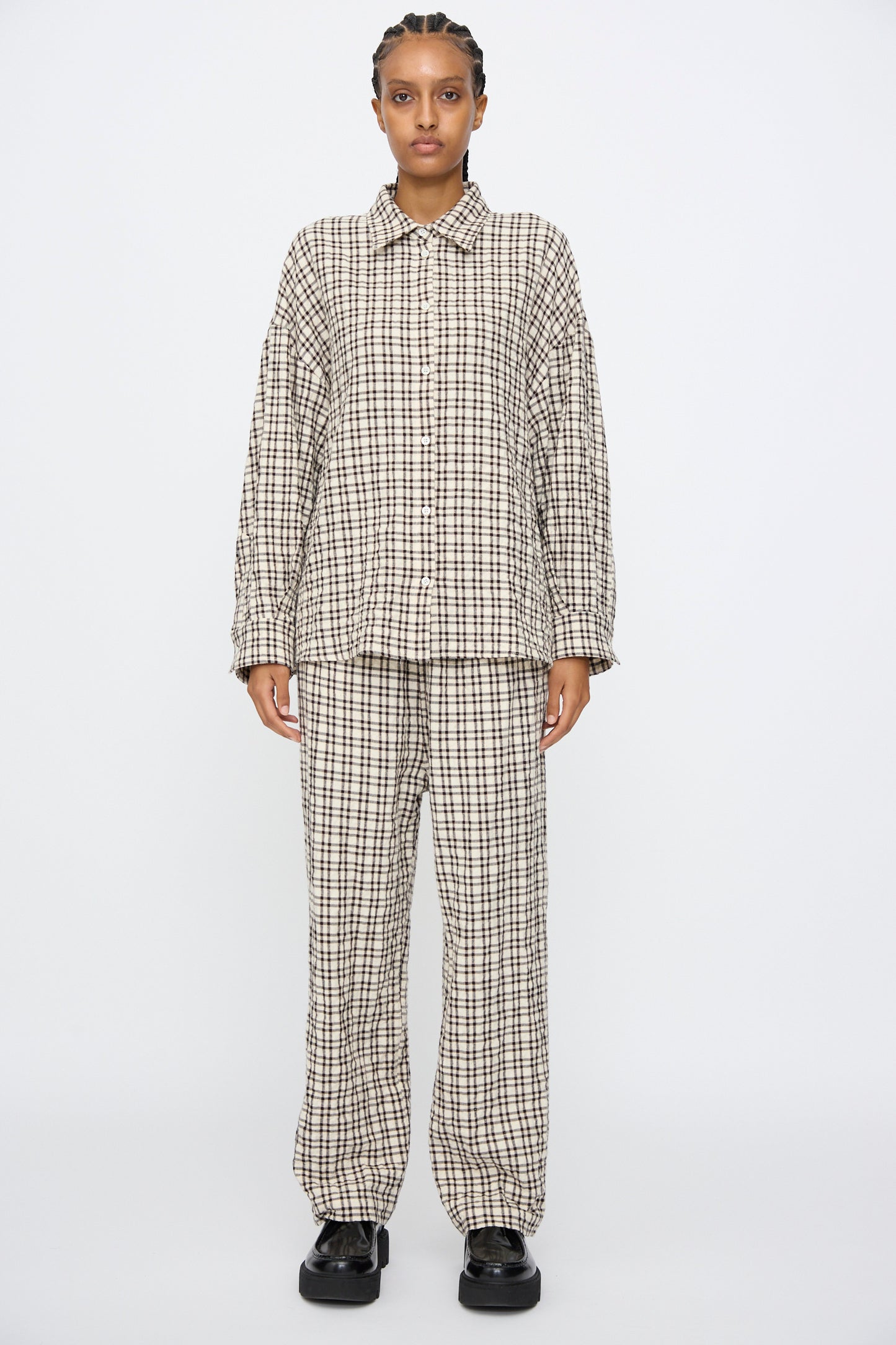 A person stands facing forward, wearing the AVN Over Shirt in White and Black Check with matching pants and black shoes against a plain white background, showcasing effortless style and Italian craftsmanship.