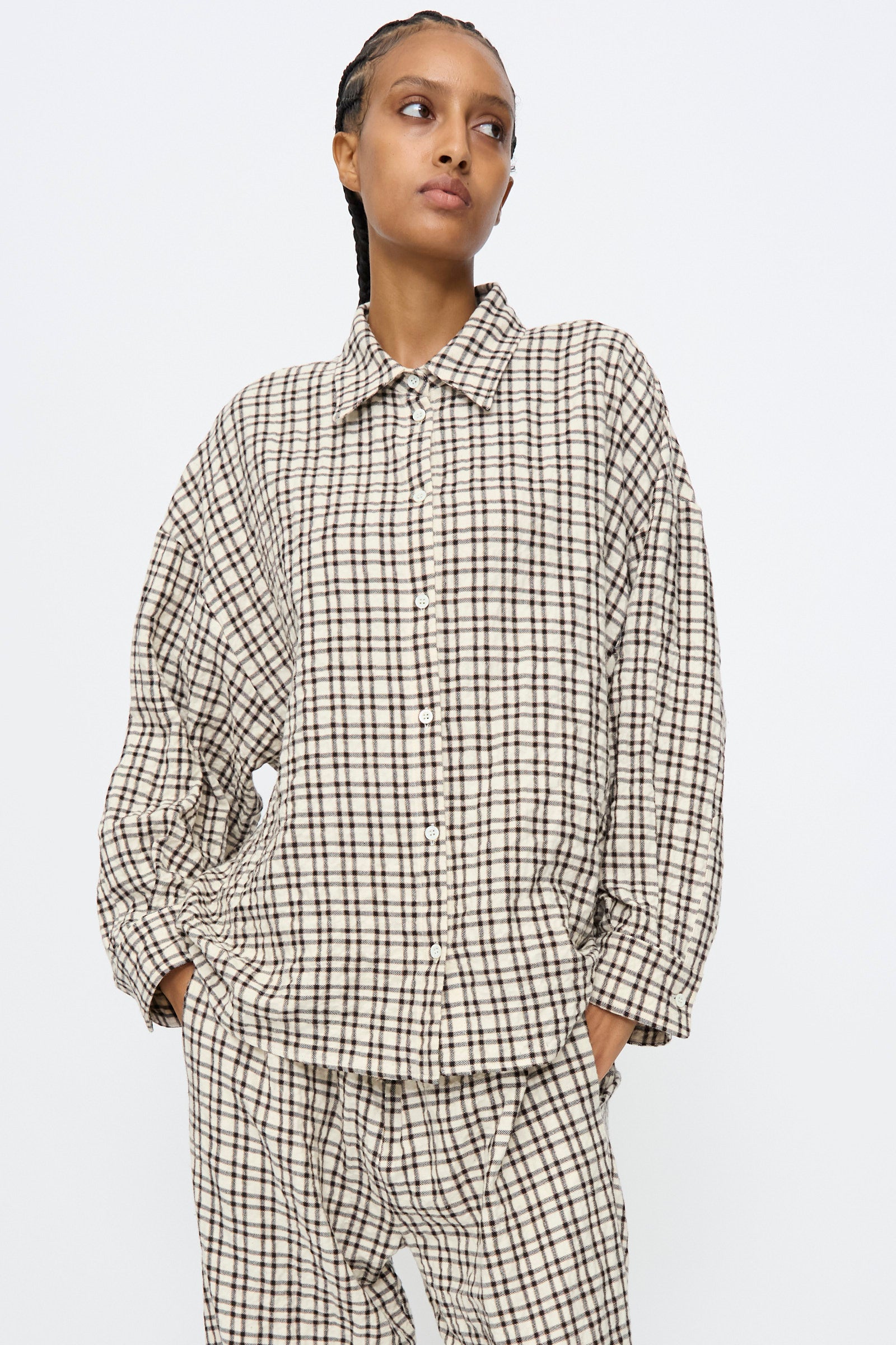 A person wears the AVN Over Shirt in White and Black Check, a loose-fitting cotton shirt with long sleeves, styled with matching plaid pants and standing against a plain white background.