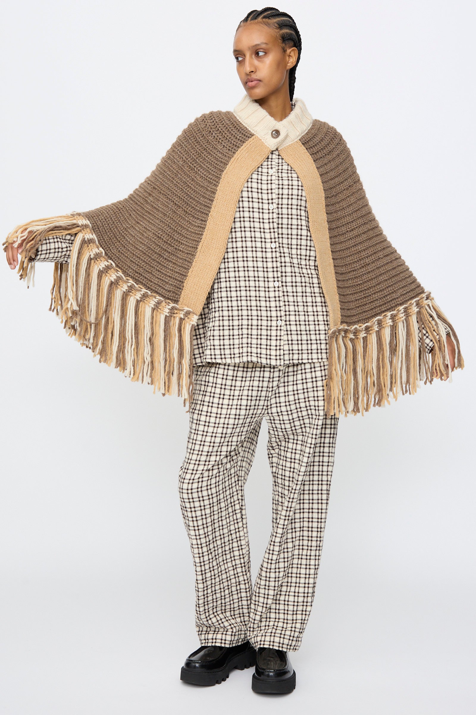 A person wearing the AVN Peru Poncho in Brown and Beige Placket stands against a plain white background, styled over a plaid shirt, pants, and black shoes.