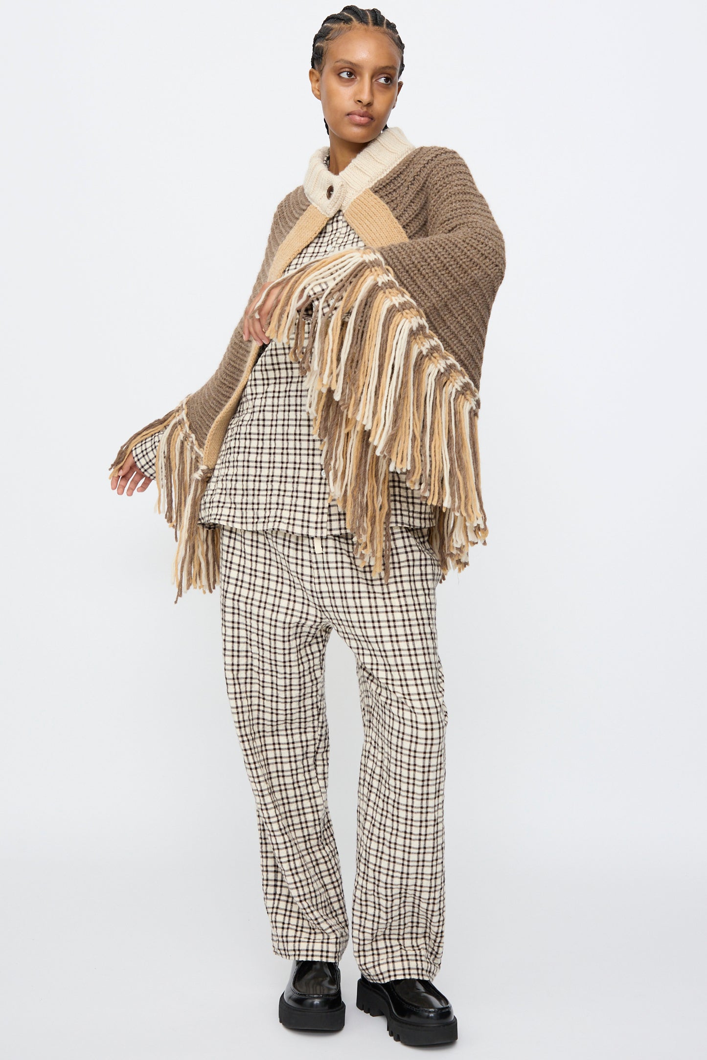 A person wears the AVN Peru Poncho in Brown and Beige Placket over a beige and black checkered outfit with black shoes, standing against a plain white background.