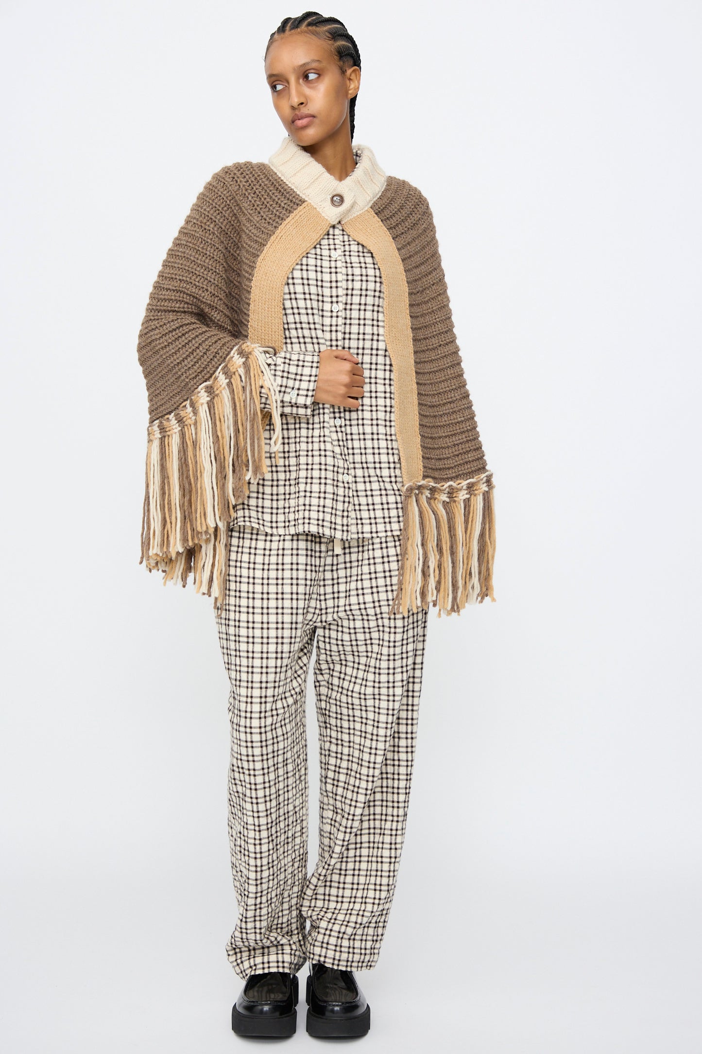 A person wears the AVN Peru Poncho in Brown and Beige Placket over a checked pajama-style shirt and pants with black shoes, standing against a plain white background.