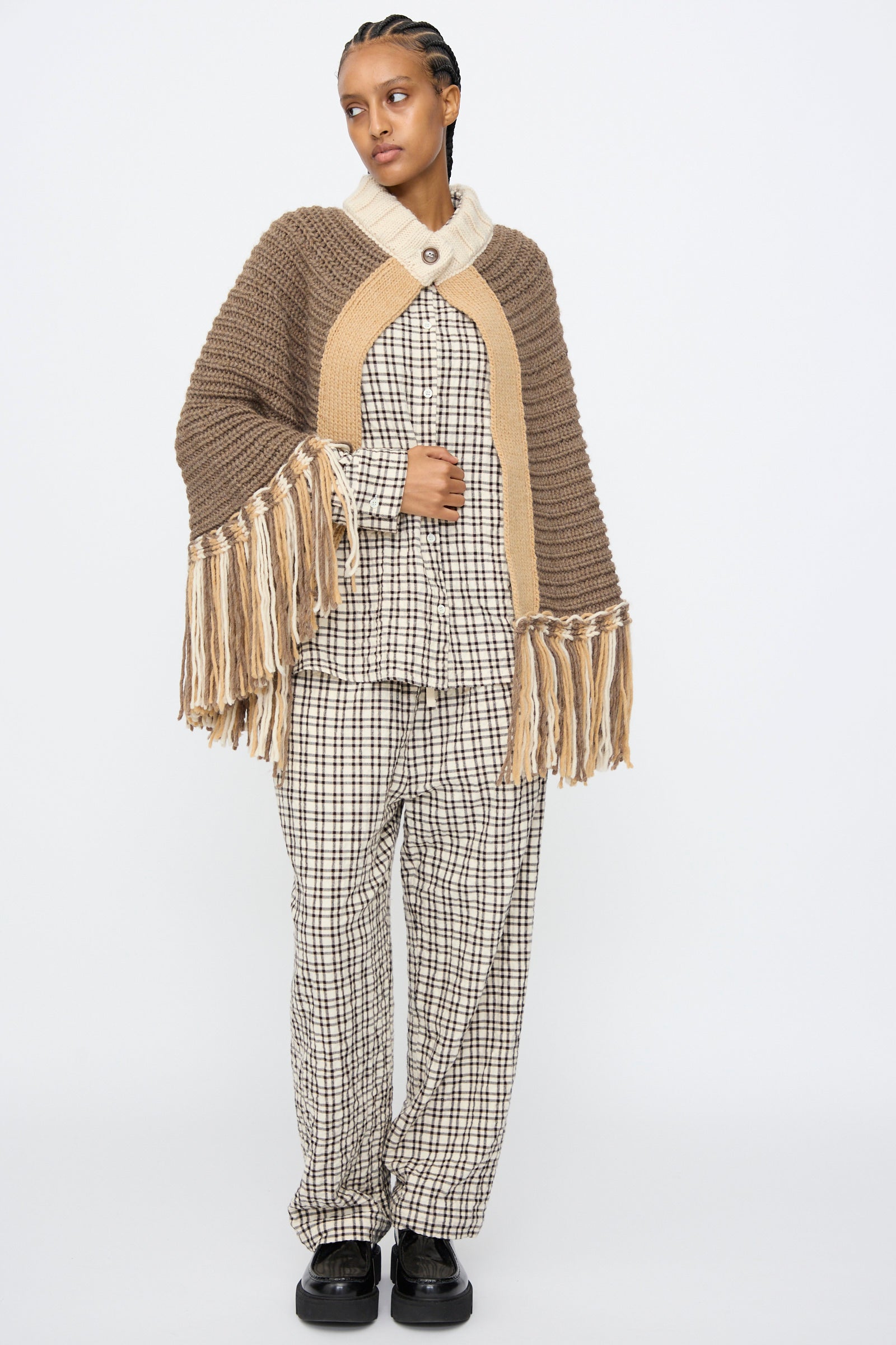 A person wears the AVN Peru Poncho in Brown and Beige Placket over a checked pajama-style shirt and pants with black shoes, standing against a plain white background.
