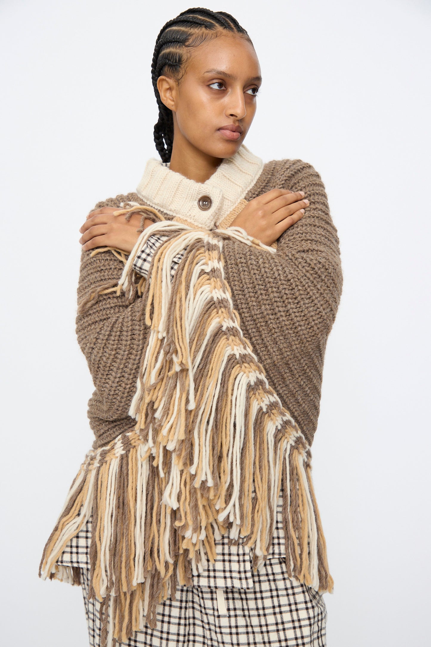 A person with braided hair crosses their arms, wearing the AVN Peru Poncho in Brown and Beige Placket over a plaid skirt against a plain background.