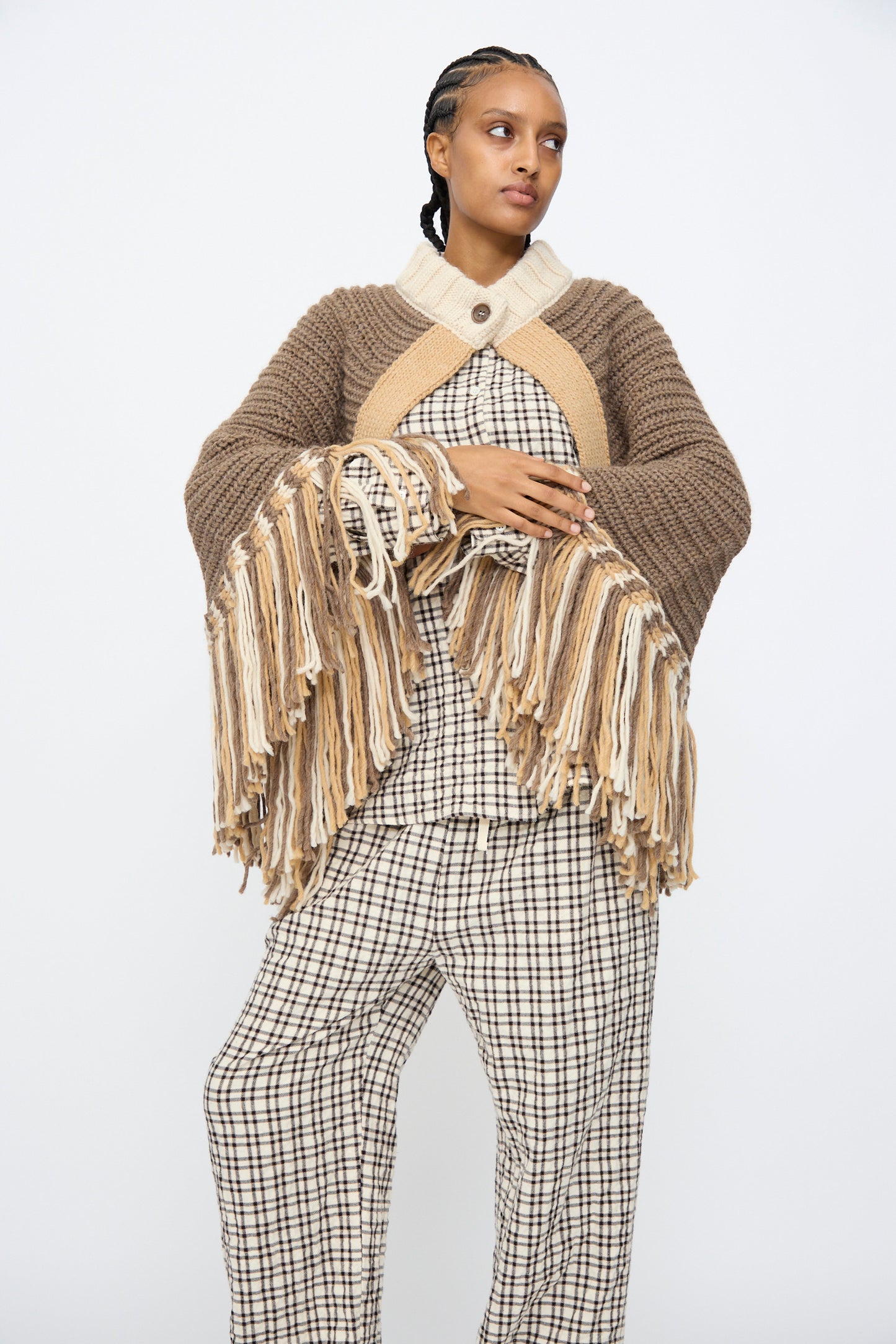 A person models the AVN Peru Poncho in Brown and Beige Placket, featuring a chunky knit with long fringe, over a checked outfit against a plain white background.