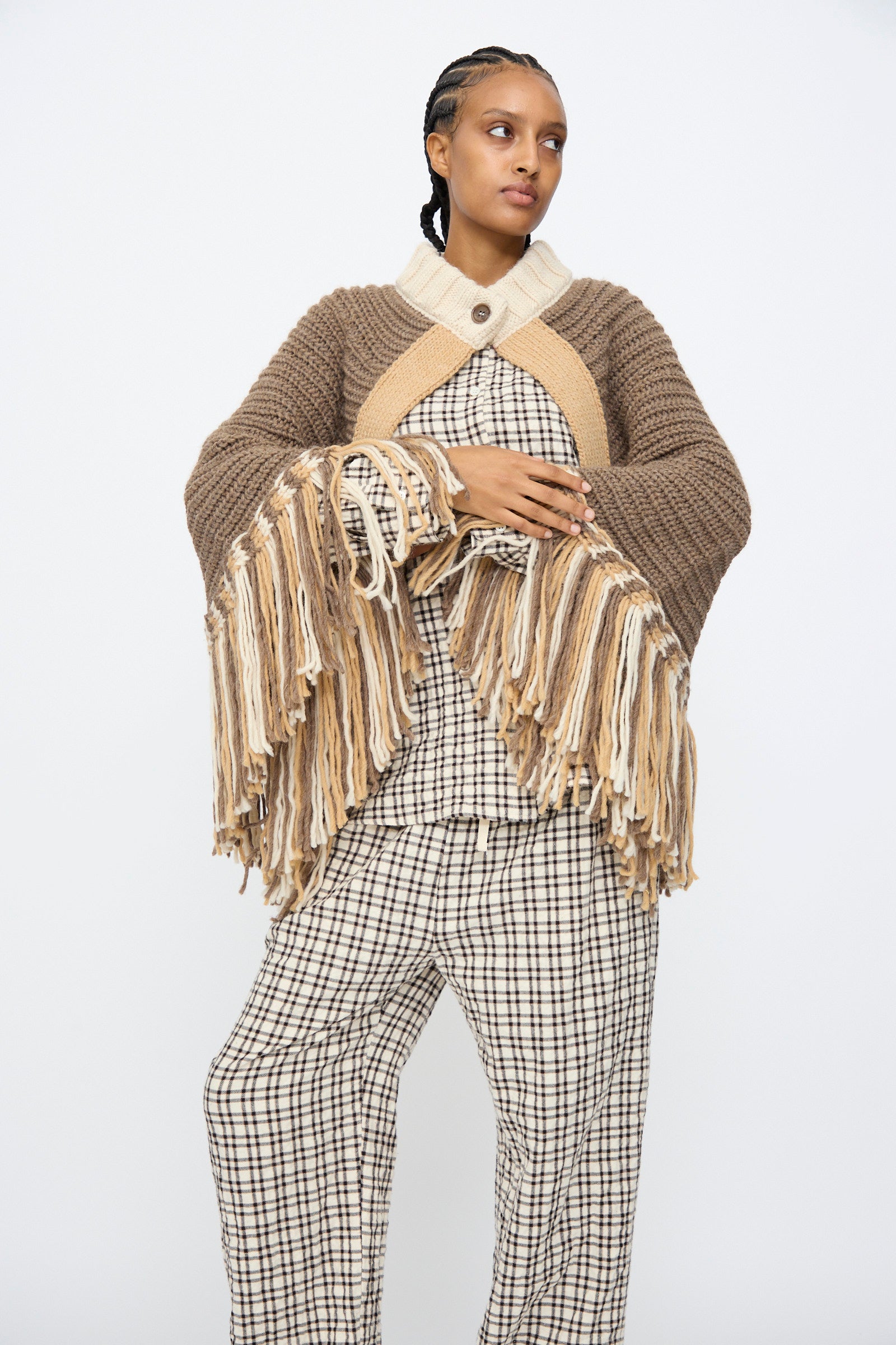 A person models the AVN Peru Poncho in Brown and Beige Placket, featuring a chunky knit with long fringe, over a checked outfit against a plain white background.