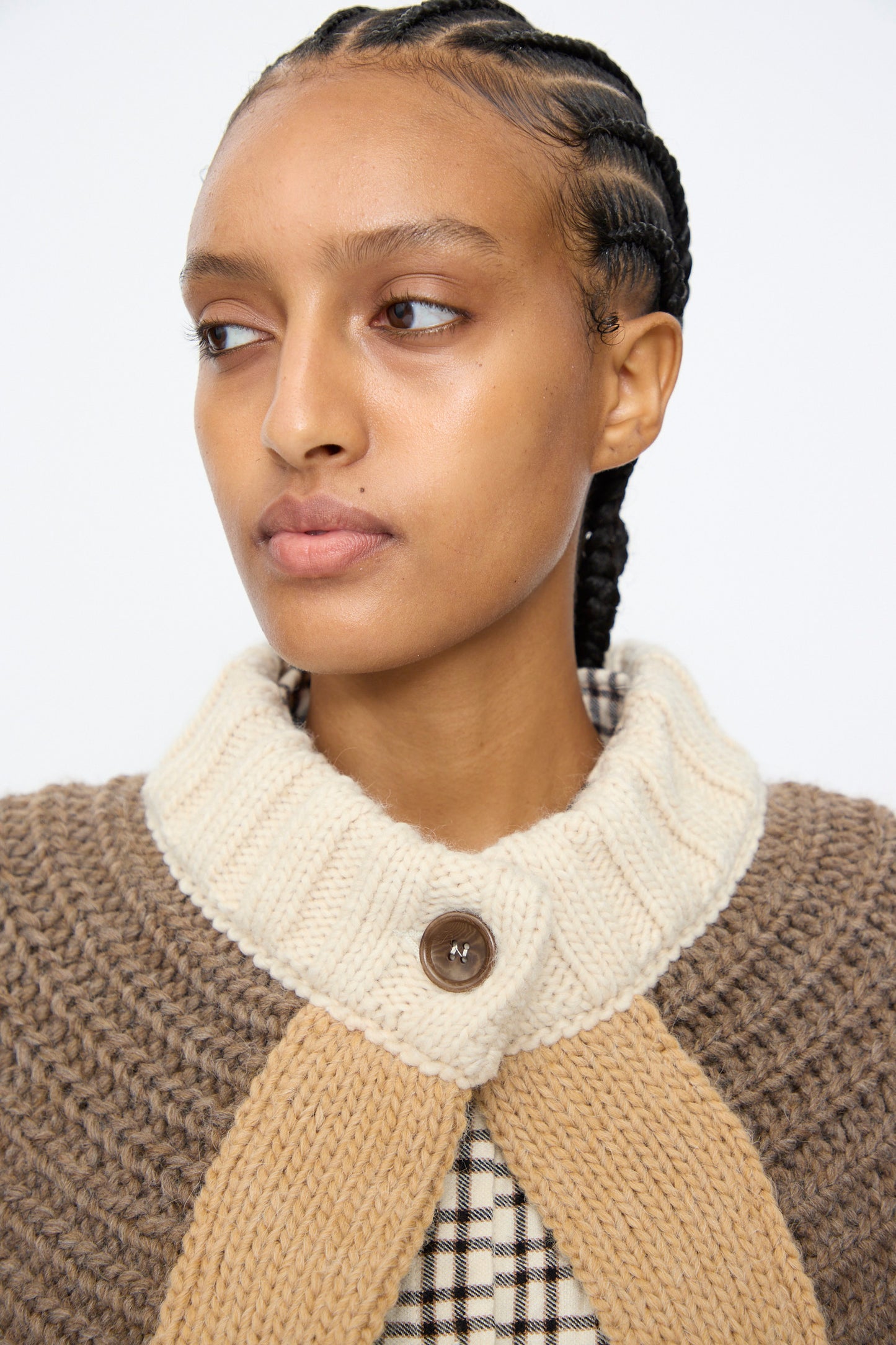 A person with braided hair wears the AVN Peru Poncho in Brown and Beige Placket over a plaid shirt, gazing slightly to the side against a plain background.