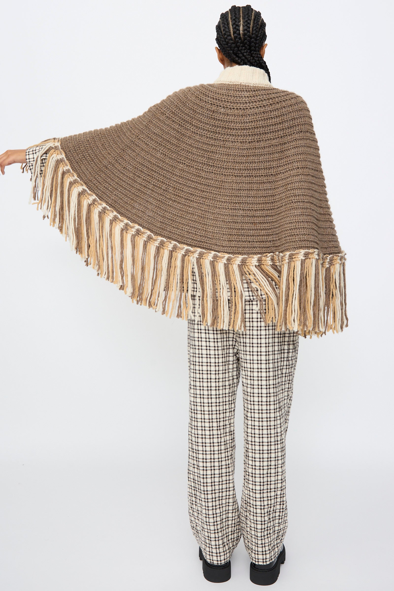 A person stands with their back to the camera wearing the AVN Peru Poncho in Brown and Beige Placket over plaid pants and black platform shoes.
