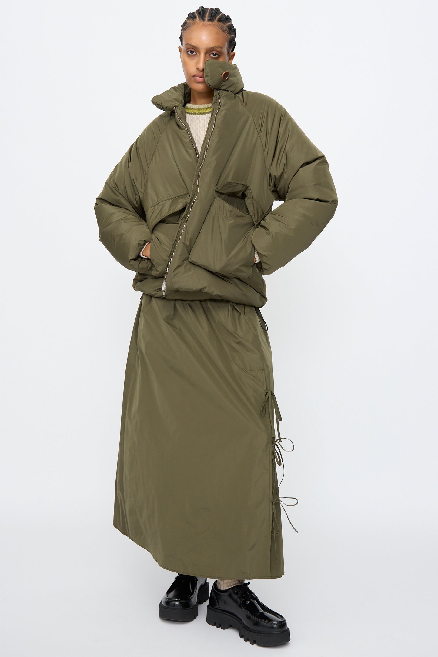 A person wears the AVN Piumino Jacket in Military Green, paired with a matching long skirt and black platform shoes, standing against a plain white background.