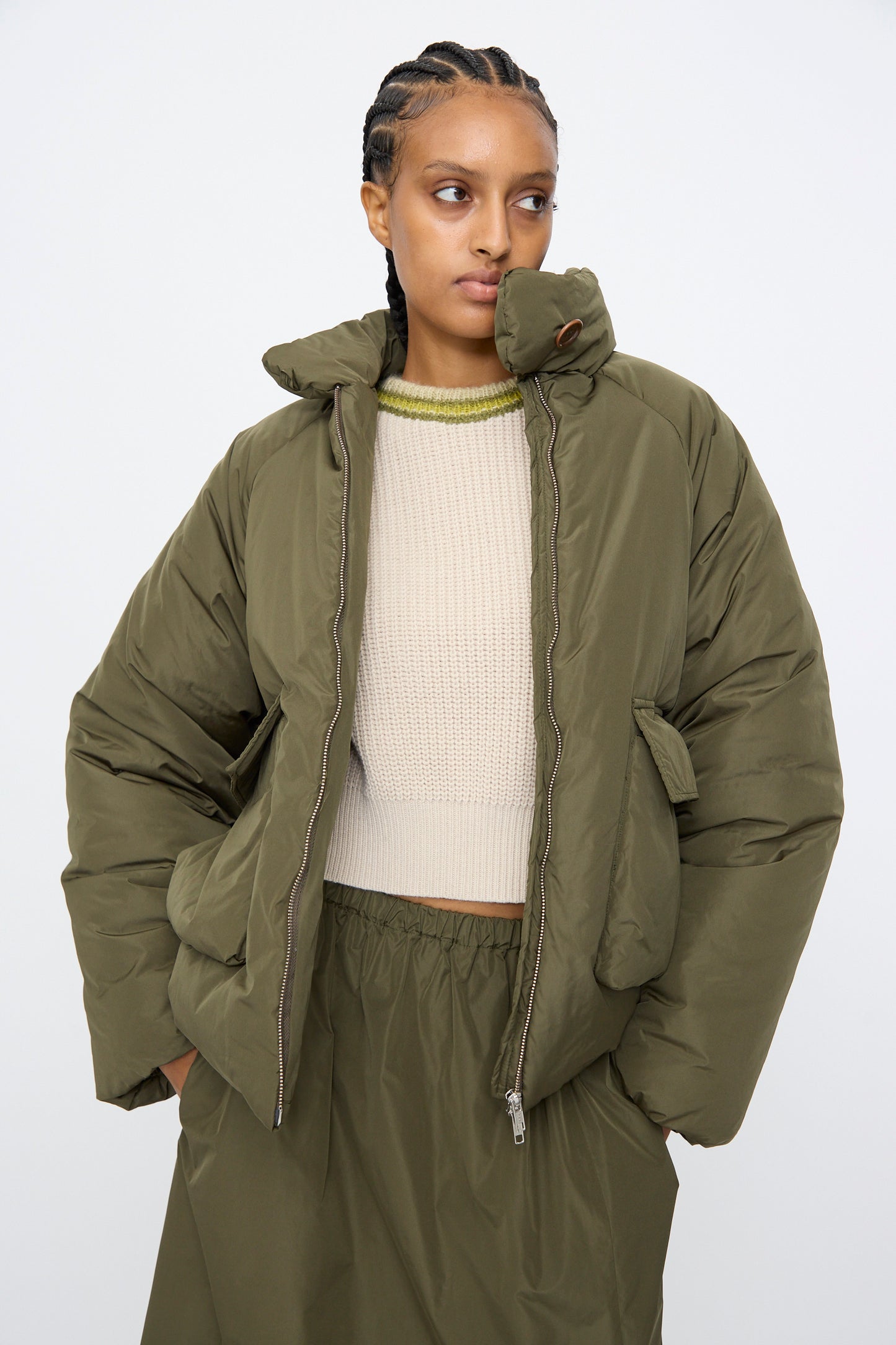 A person wears the AVN Piumino Jacket in Military Green, styled oversized over a beige knit sweater and coordinating olive green skirt, standing against a plain light background.