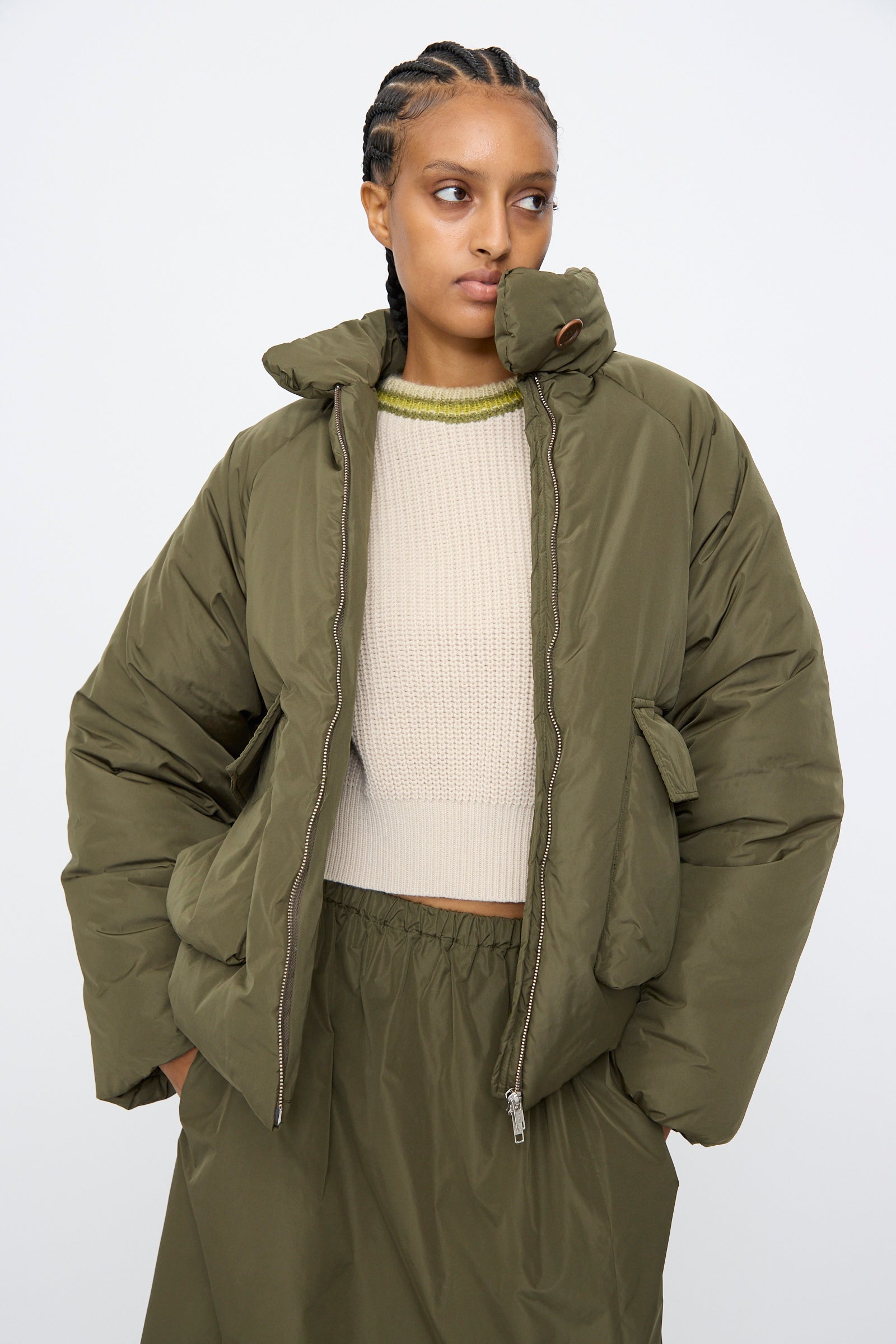 A person wears the AVN Piumino Jacket in Military Green, styled oversized over a beige knit sweater and coordinating olive green skirt, standing against a plain light background.