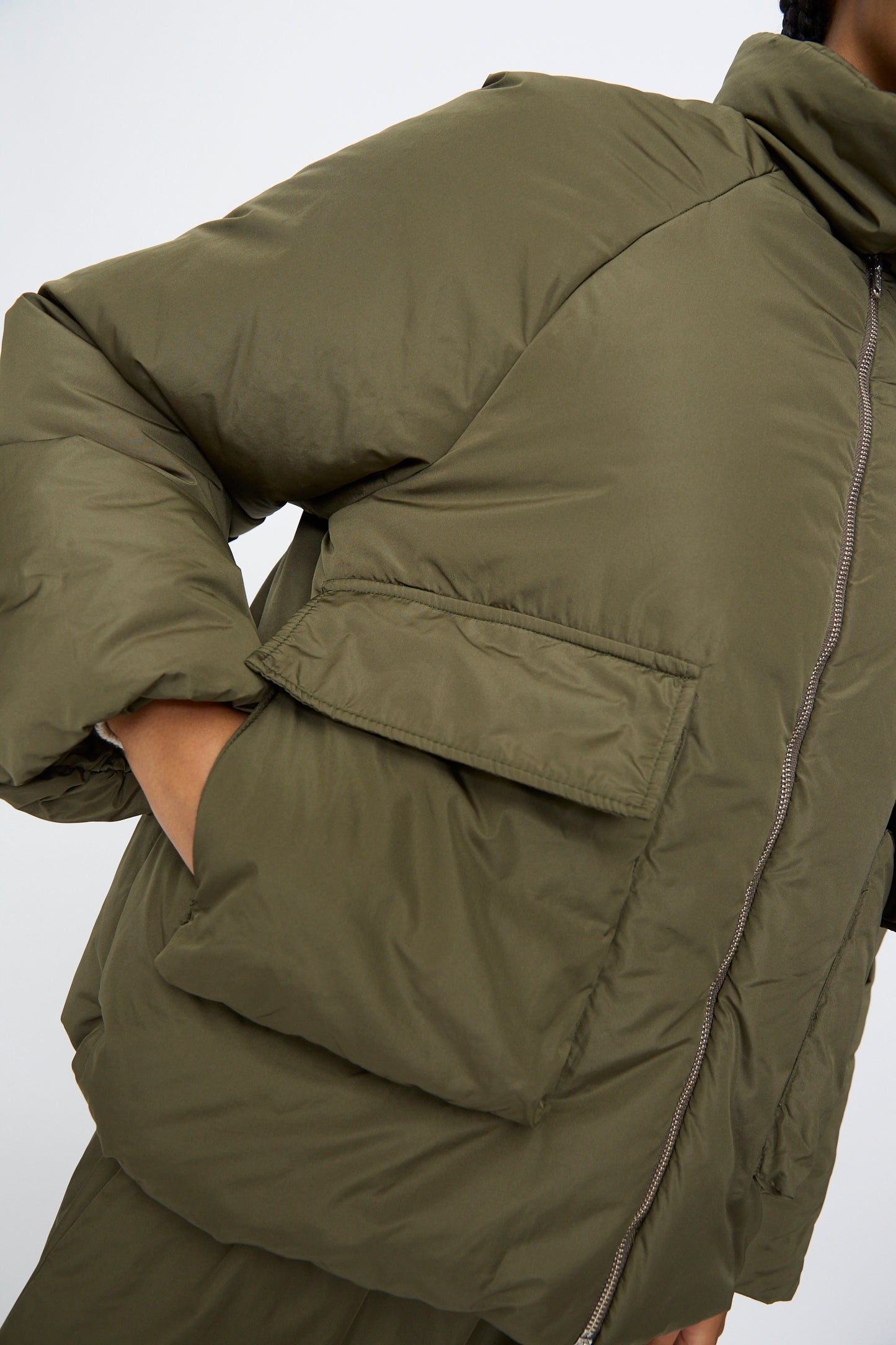 A person wears the AVN Piumino Jacket in Military Green—an oversized puffer with a large front pocket and zipper, hand partly in the pocket, set against a plain background.