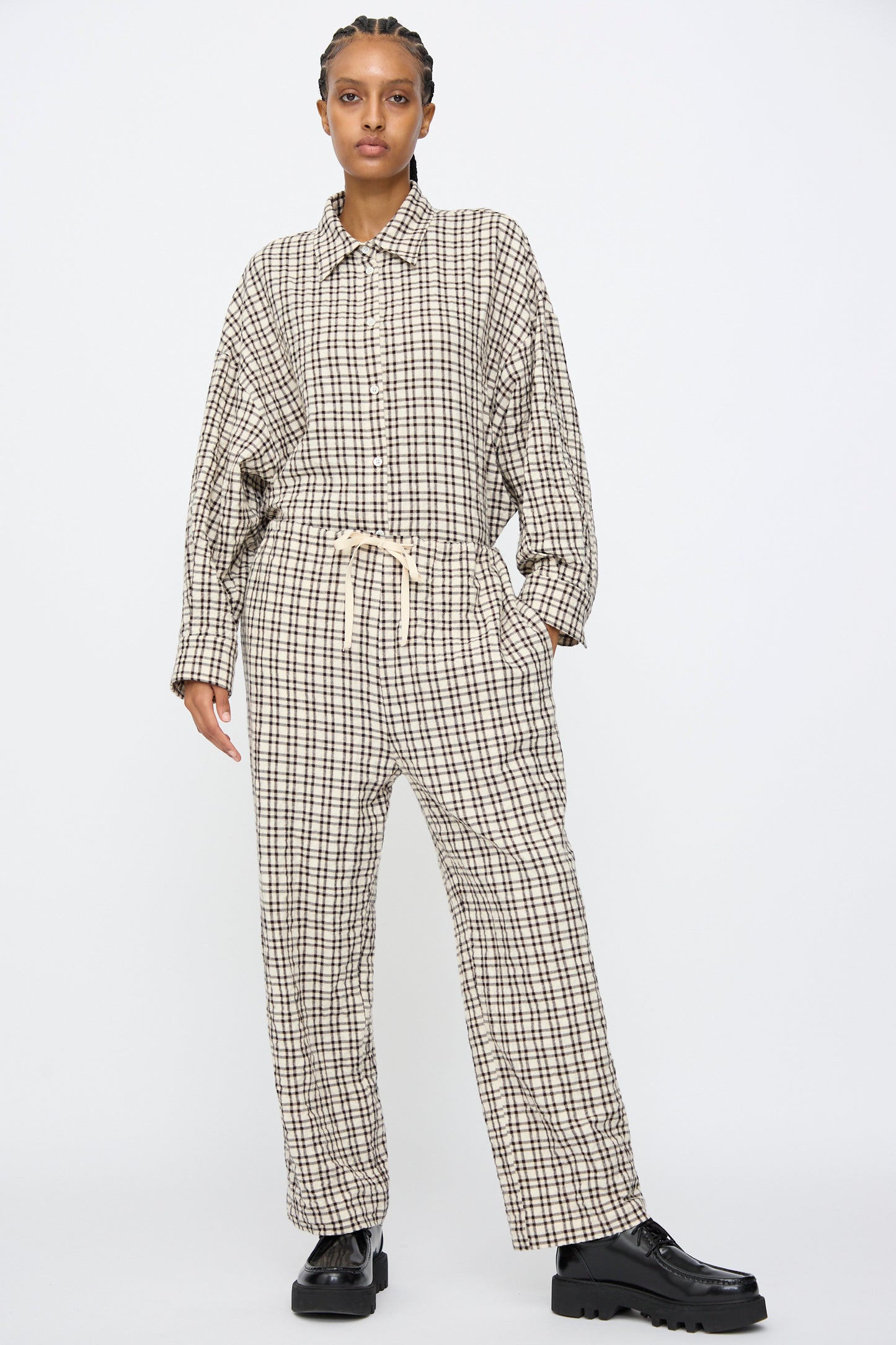 A person stands against a plain background wearing AVN's Pj Pants in White and Black Check—relaxed fit, checkered cotton PJ style—paired with a loose plaid button-up and black chunky shoes.