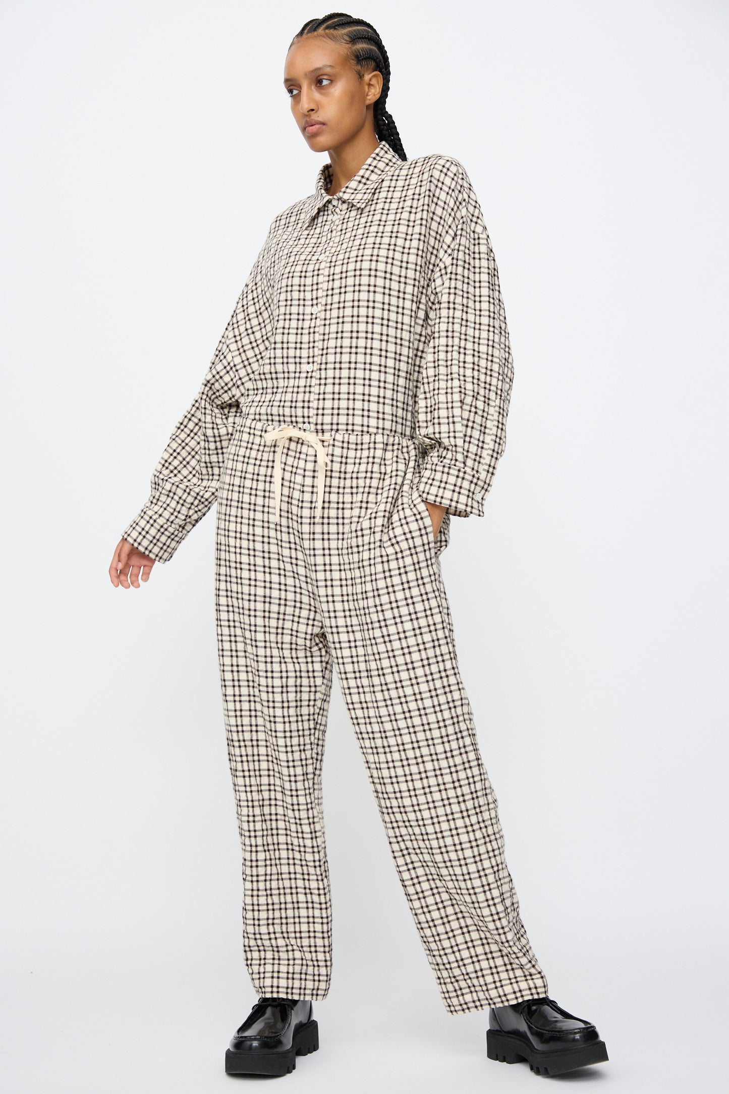 A person wears AVN's Pj Pants in White and Black Check paired with a loose beige and black checkered shirt and black shoes, standing against a plain white background.