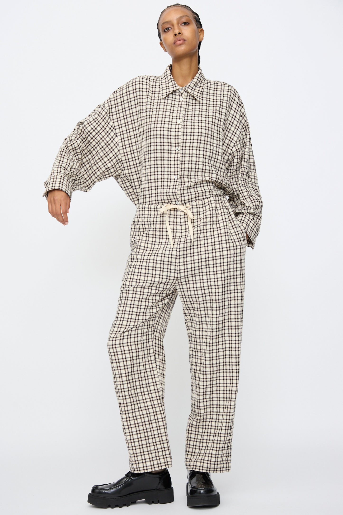 A person wears AVN Pj Pants in White and Black Check—relaxed fit, cotton, checkered style—with a matching oversized shirt and black platform shoes, standing against a plain white background.