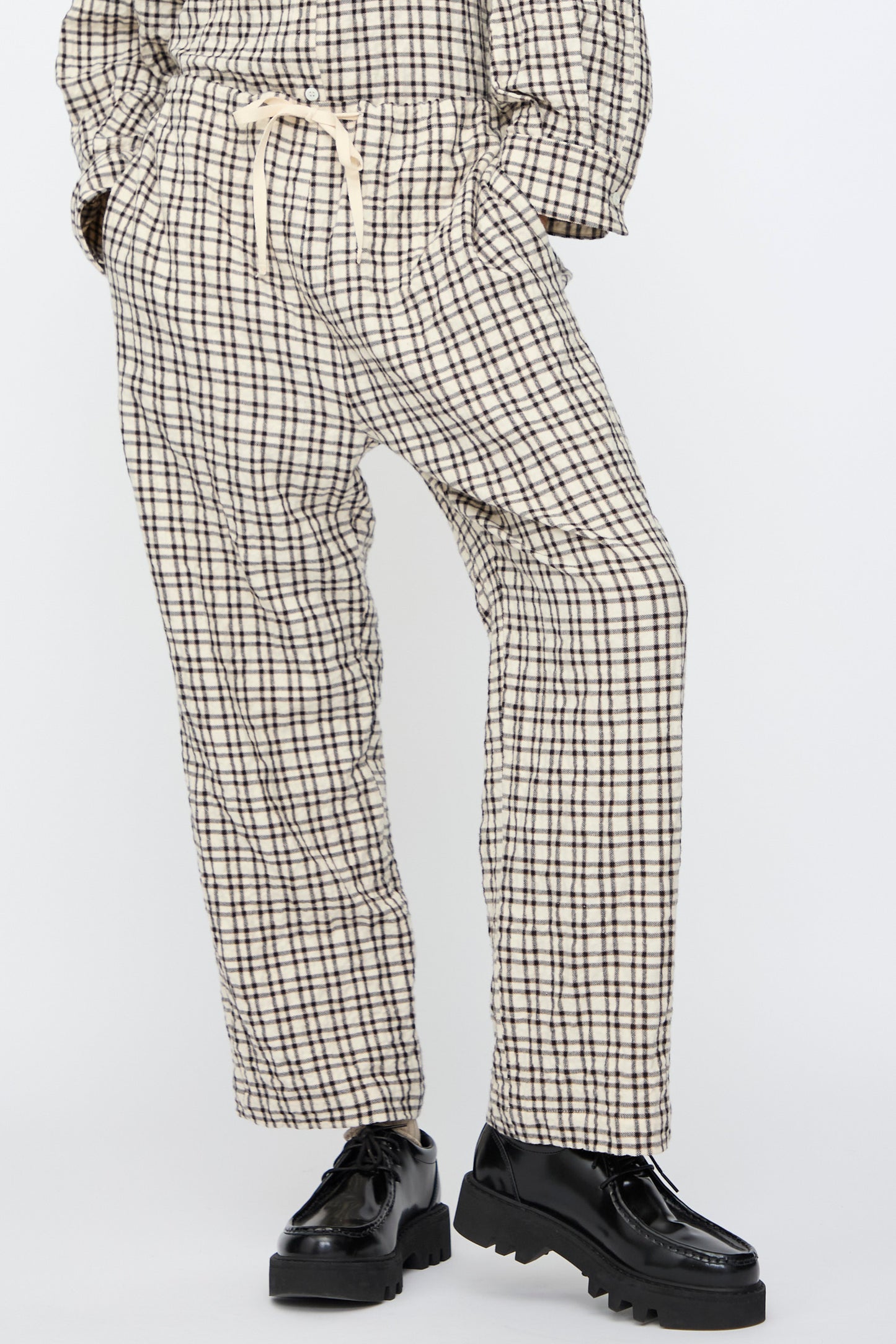 A person wears AVN’s Pj Pants in White and Black Check—relaxed fit, drawstring waist—paired with black chunky shoes, standing against a plain light background.