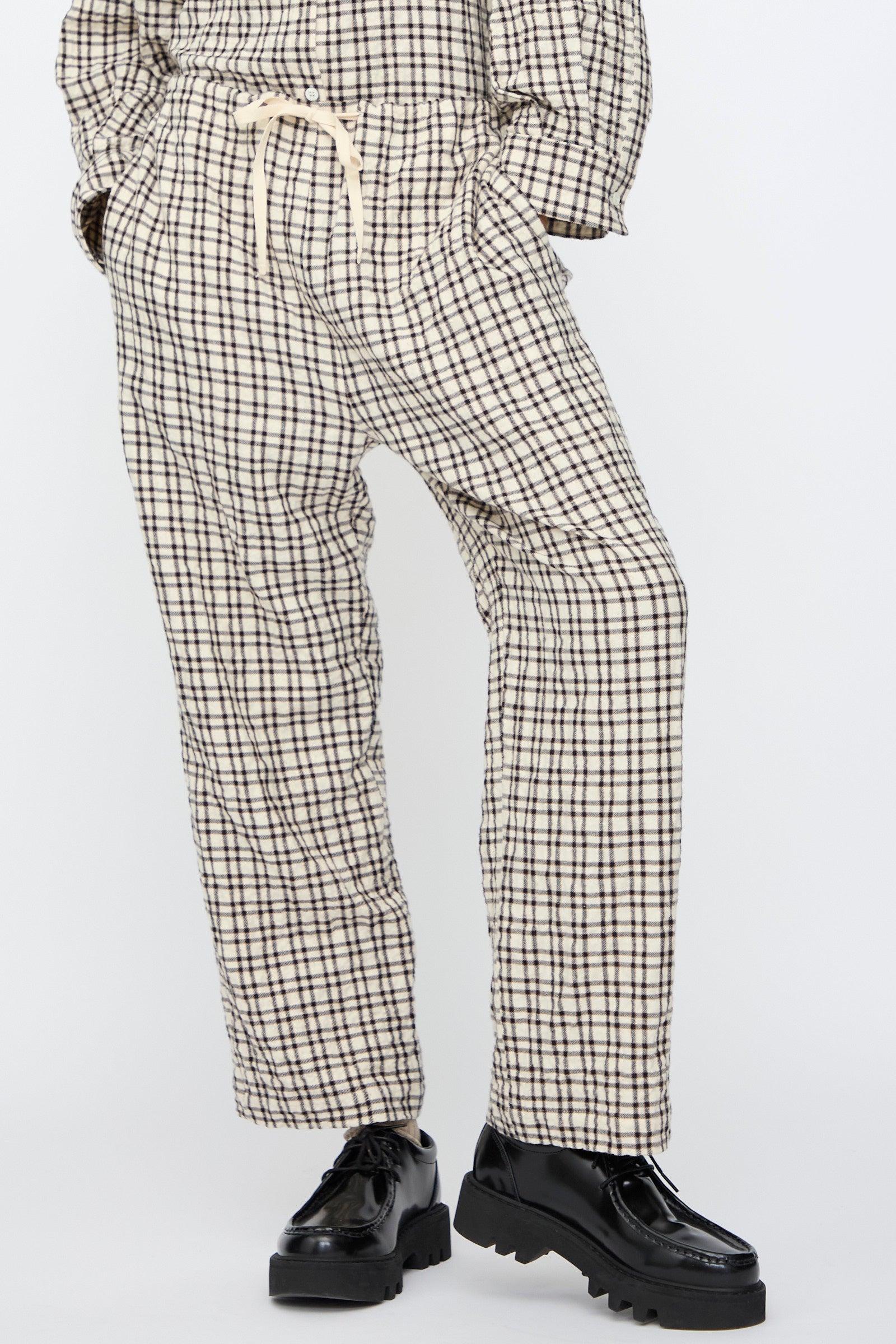 A person wears AVN’s Pj Pants in White and Black Check—relaxed fit, drawstring waist—paired with black chunky shoes, standing against a plain light background.