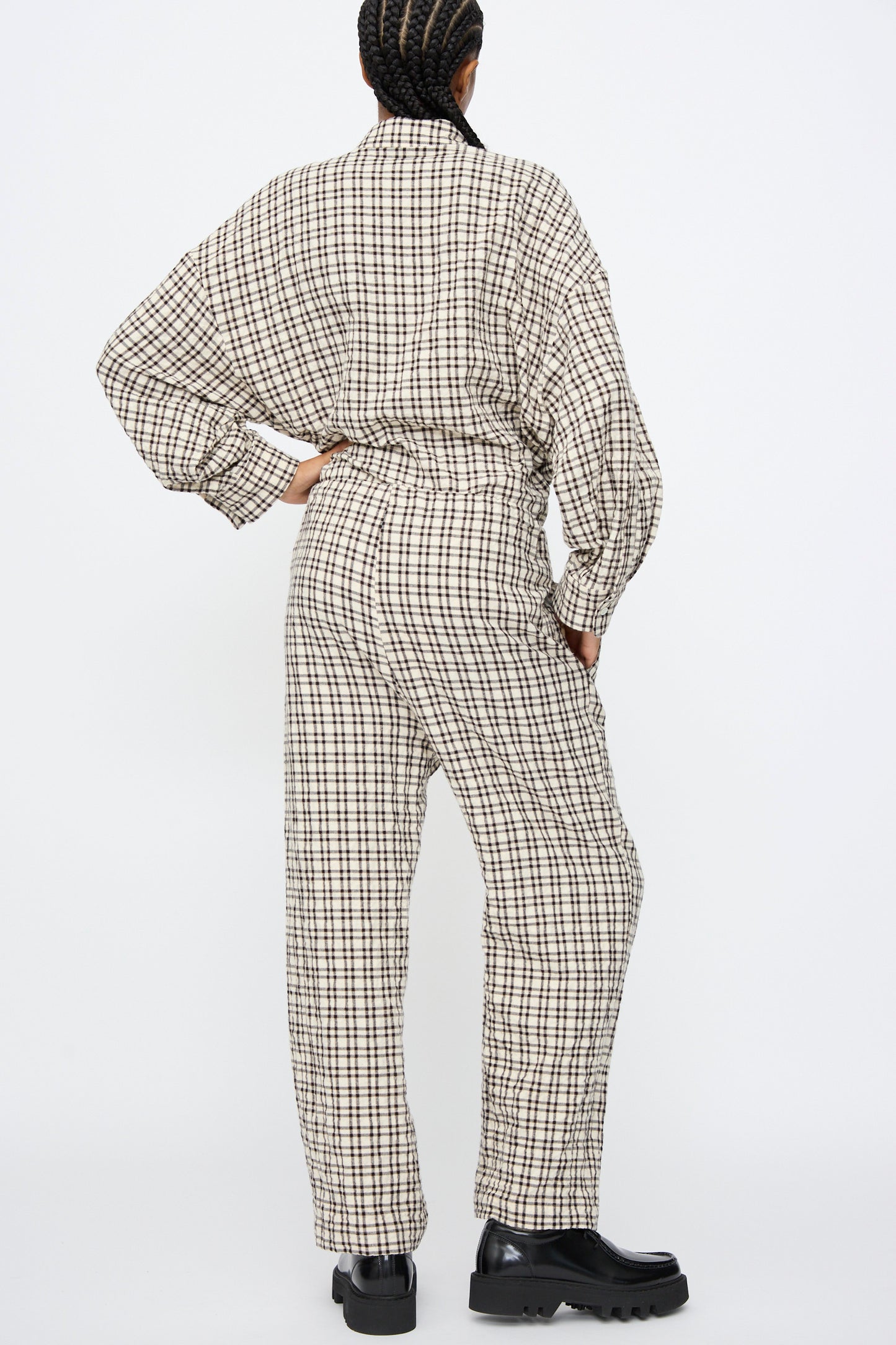Person stands with back to camera, wearing AVN Pj Pants in White and Black Check and black chunky shoes against a plain white background.