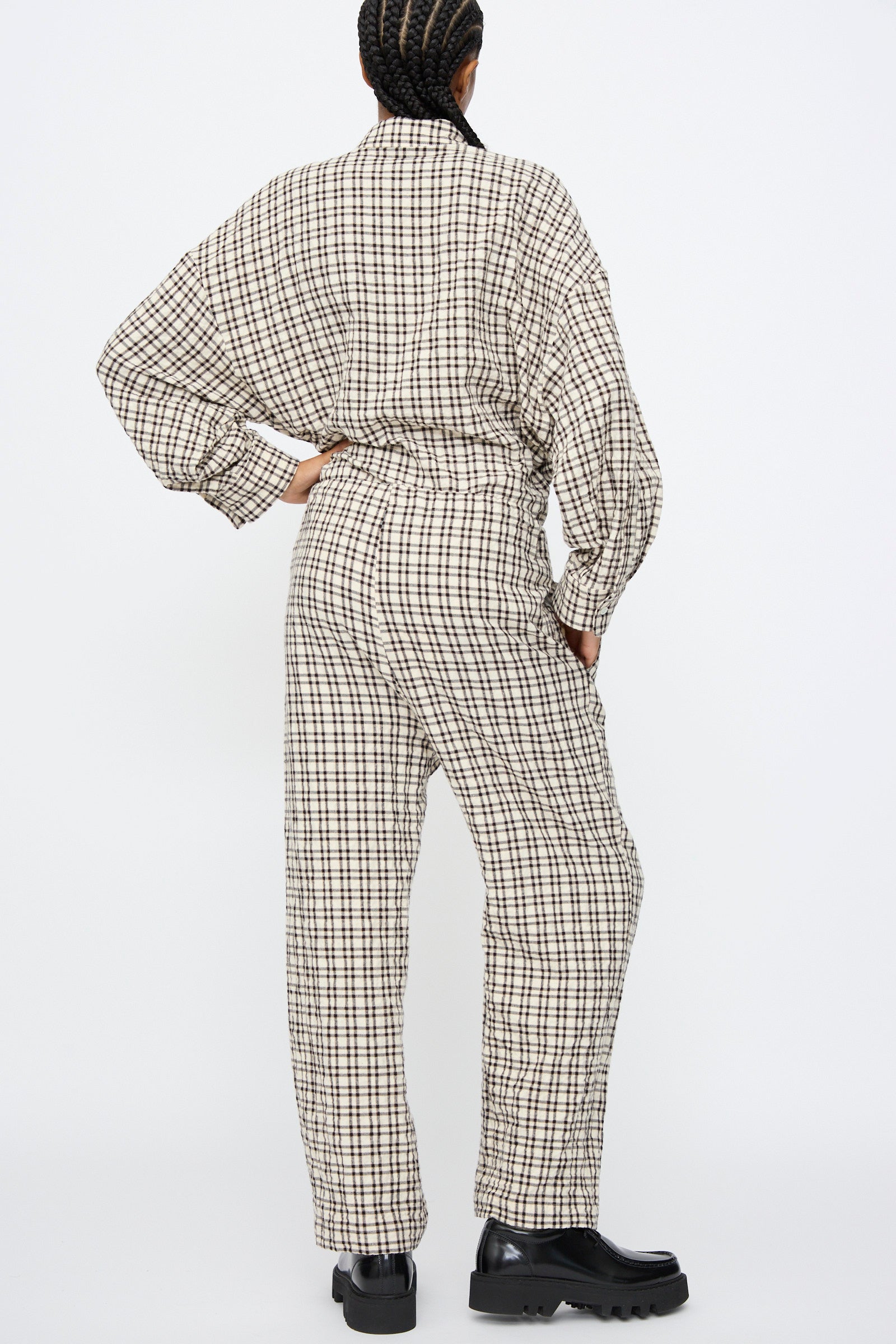 Person stands with back to camera, wearing AVN Pj Pants in White and Black Check and black chunky shoes against a plain white background.
