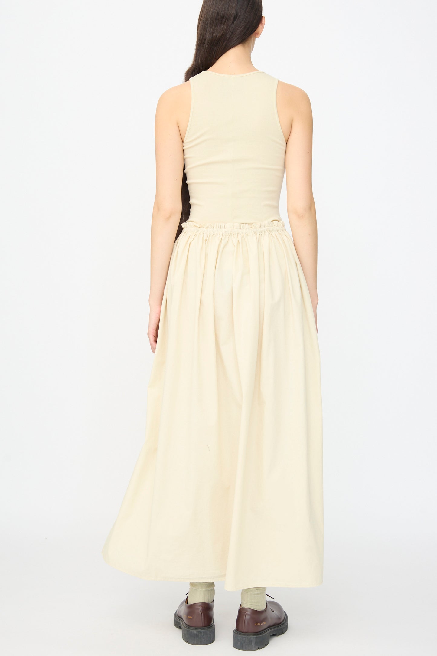 A person faces away wearing the AVN Tank Dress in Sand, a sleeveless beige cotton piece styled with brown shoes against a plain white background.