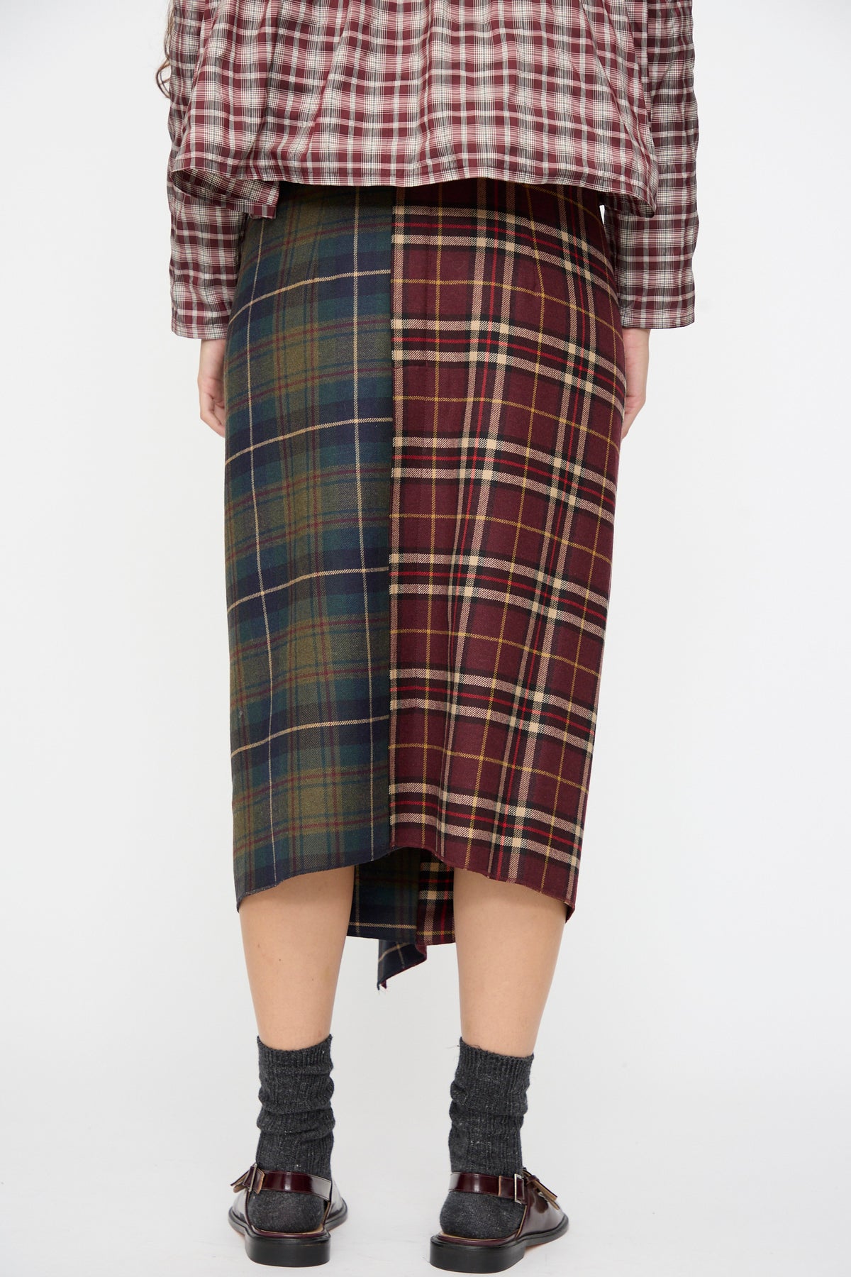 Back view of an AVN Volant Skirt in Bordeaux and Military Green Check, a color-blocked plaid wool midi skirt worn with a matching top, dark socks, and buckled shoes.