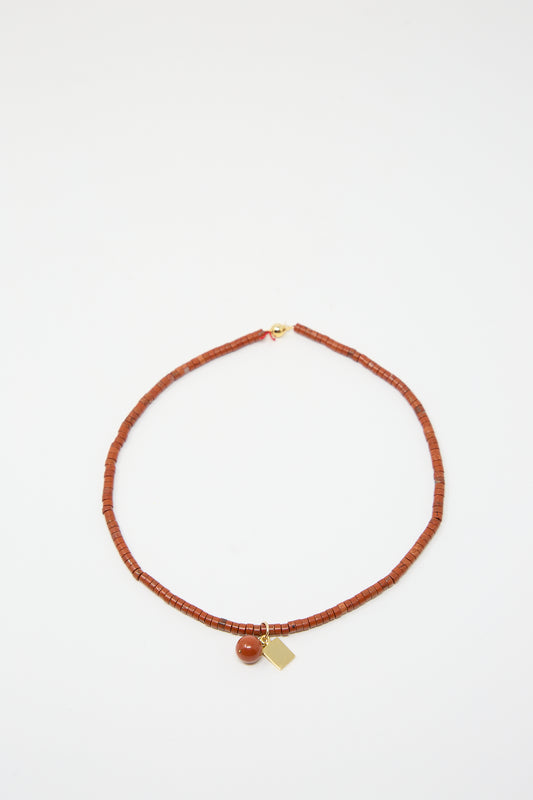 A beaded necklace featuring Red Jasper Heishi beads, a small gold tag, and a round pendant, displayed on a plain white background. Handmade in NYC. This is the 14K Gold Plated Brass Heishi Necklace in Red Jasper by Abby Carnevale.