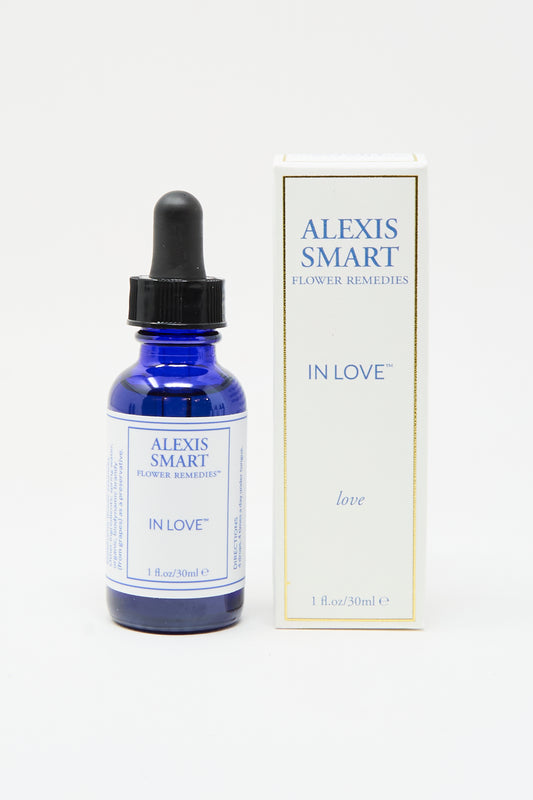 A blue glass dropper bottle labeled "Alexis Smart Flower Remedies - In Love" stands next to its matching white and gold box, both displaying the brand's elegant logo. This flower essence blend supports self-love and healthy relationships.