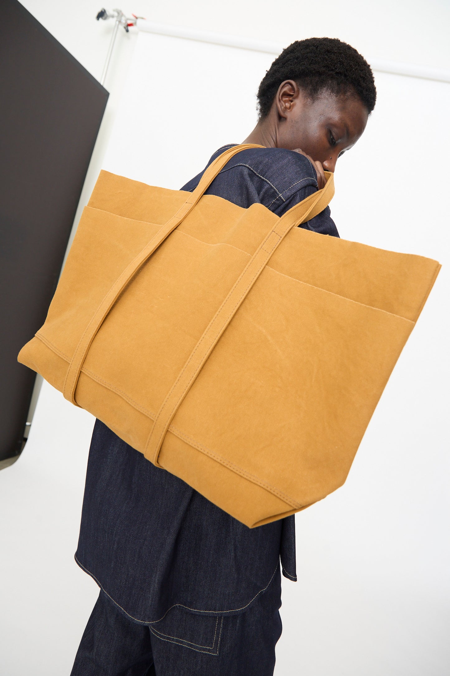 A person in dark denim carries the Amiacalva Washed Canvas 6 Pockets Large Tote in Mustard, made in Japan, over their shoulder while standing in a minimalist studio.