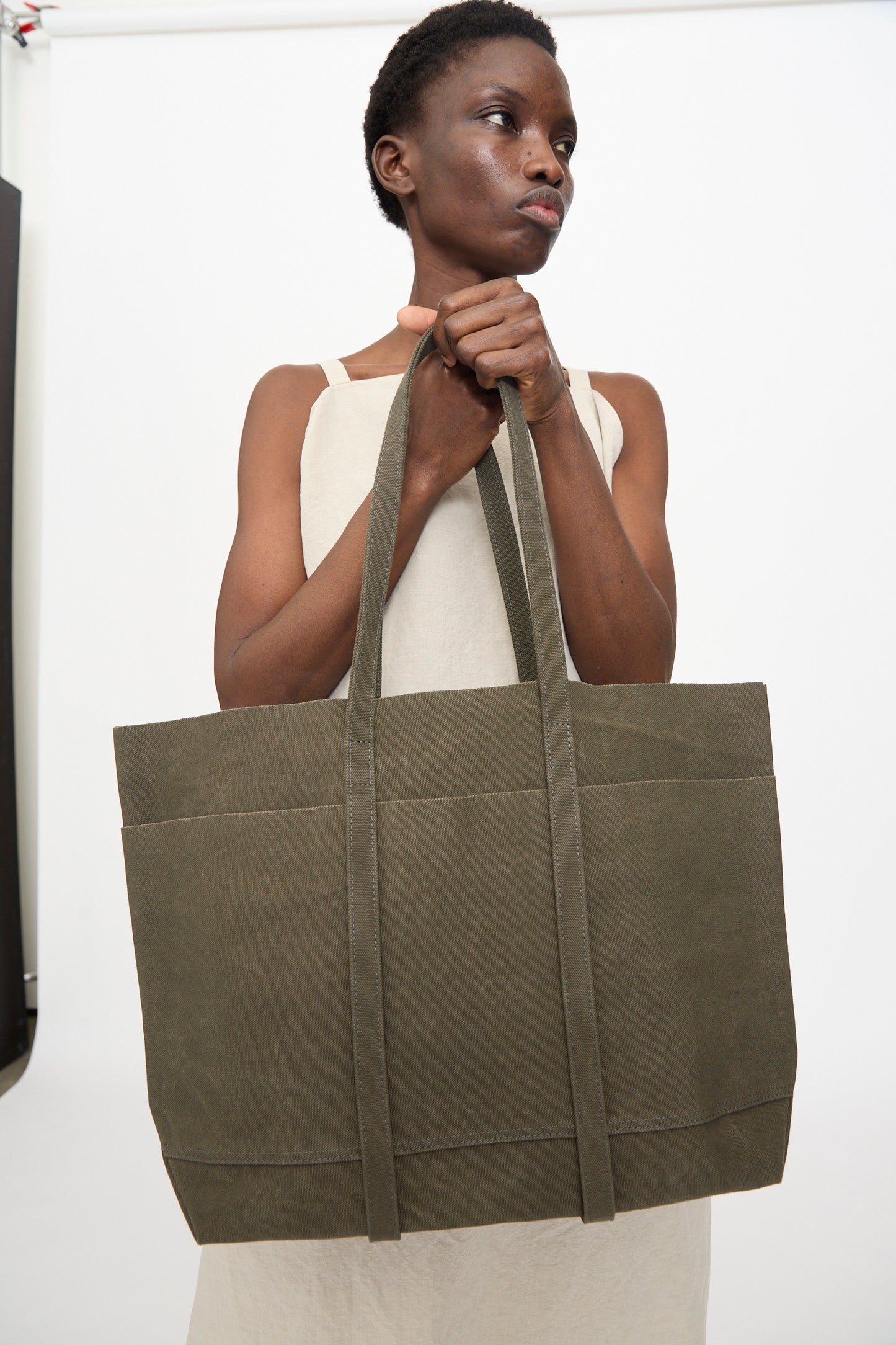 A person in a sleeveless beige dress holds the Amiacalva Washed Canvas 6 Pockets Medium Tote in Olive, made in Japan, against a plain white background.