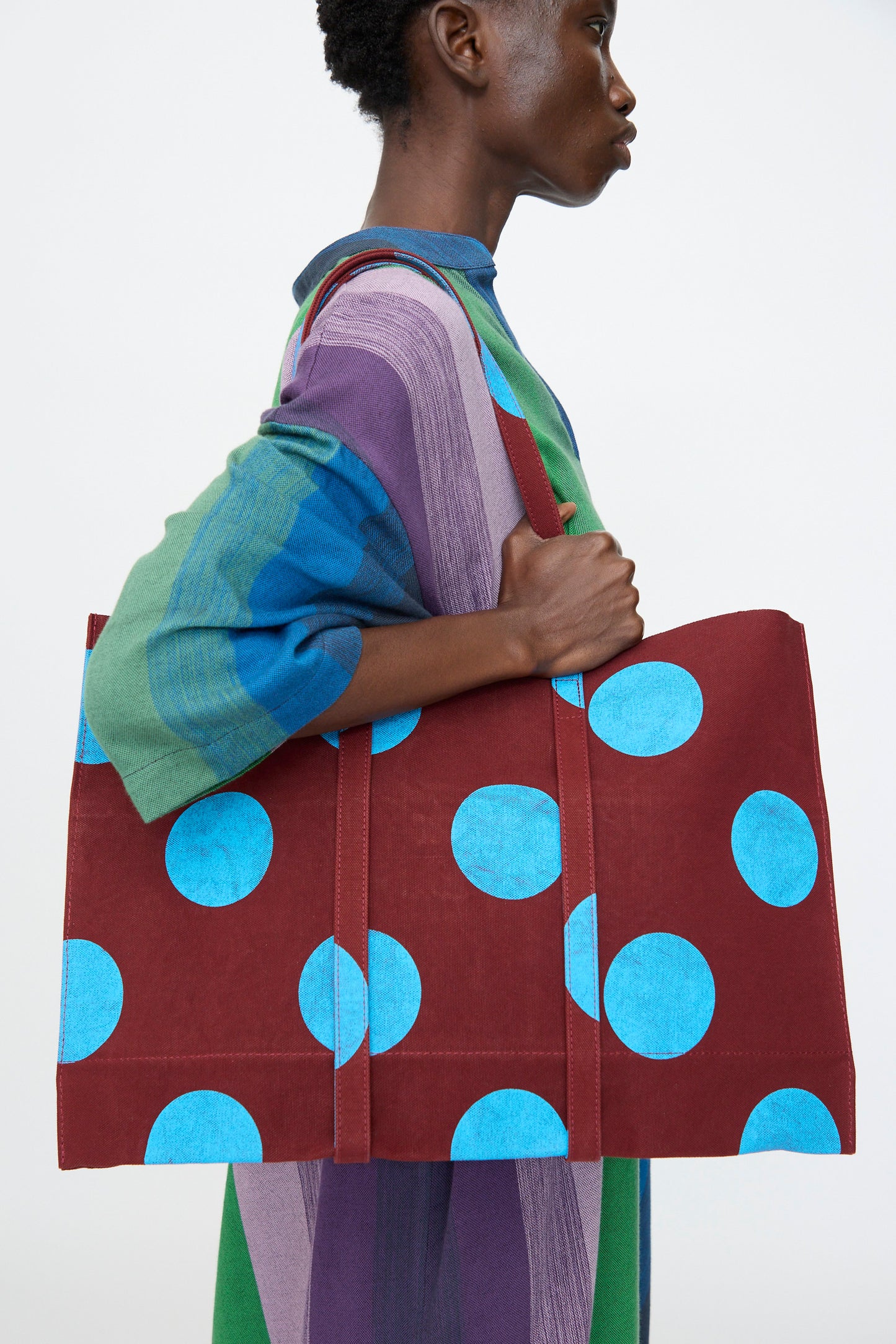 A person carries the Amiacalva Washed Canvas Medium Dot Tote in Sky Blue over their shoulder, adding a playful touch to their striped multicolored dress against a plain background.