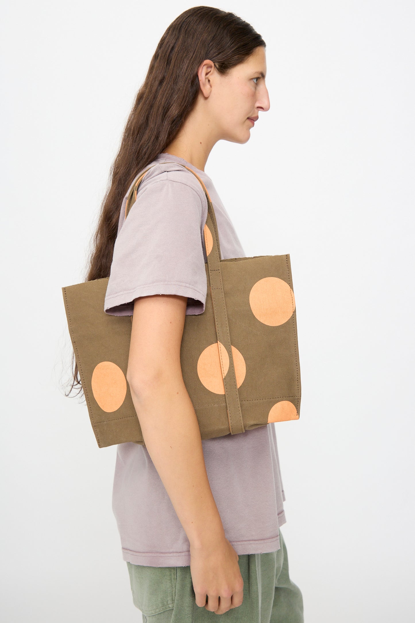 A person in a light purple shirt carries the Amiacalva Washed Canvas Small Dot Tote in Orange, made in Japan, as they stand against a plain white background.