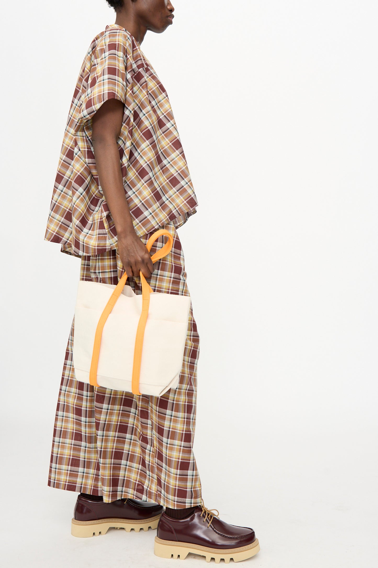 A person in a loose brown plaid outfit and chunky brown shoes holds the Amiacalva Washed Canvas Small Tote 2 Tone in Orange, made in Japan, with yellow handles, against a plain white background.