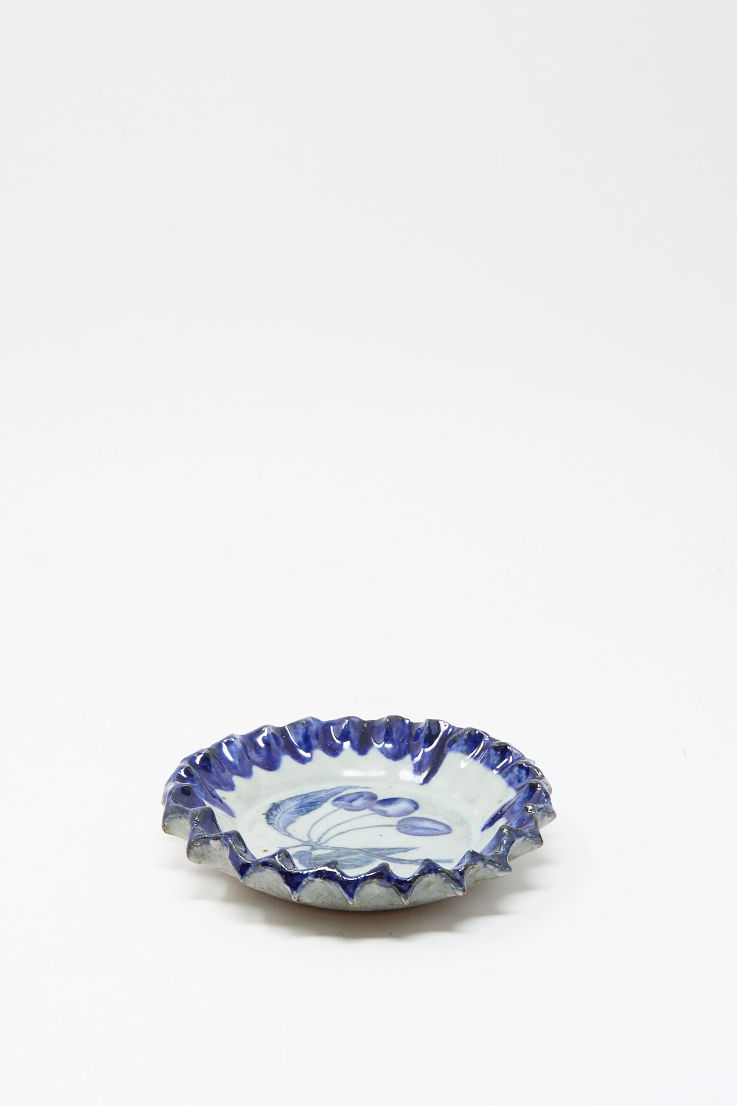 The Rustic Cherries Plate by Ariel Clute is a small, round ceramic dish with a ruffled edge and blue glaze accents on a white background, handmade in California.