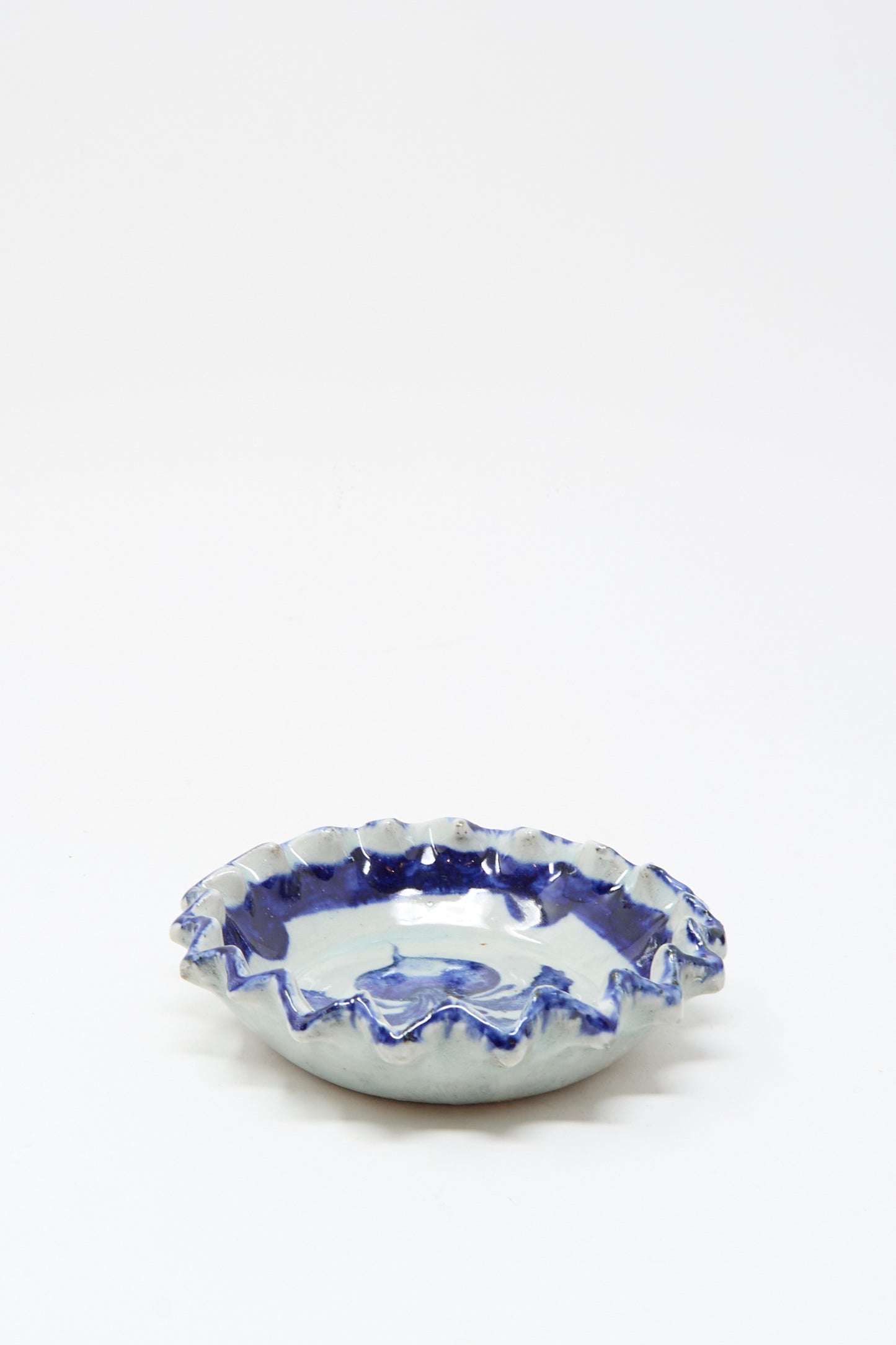The Rustic Turnip Bowl by Ariel Clute is a shallow ceramic dish with a wavy ruffled edge and blue-and-white glaze, featuring a hand-painted porcelain plate style, shown on a plain white background.