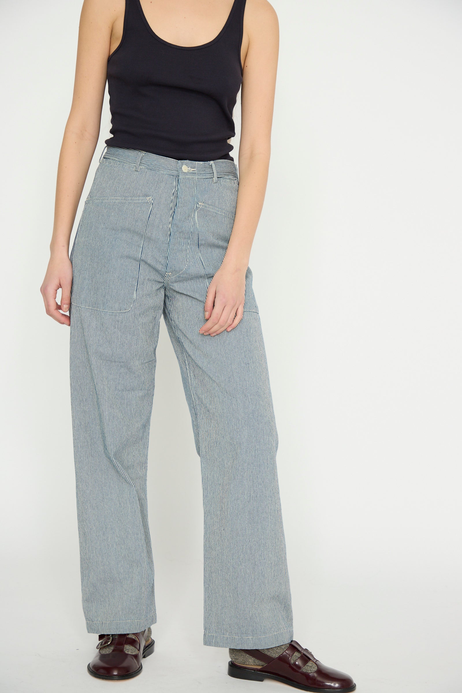 A model in a black tank top and As Ever's Brancusi Pant in Engineer Stripe, featuring a high-waisted, wide-leg silhouette, complemented by brown shoes, stands against a plain white background.