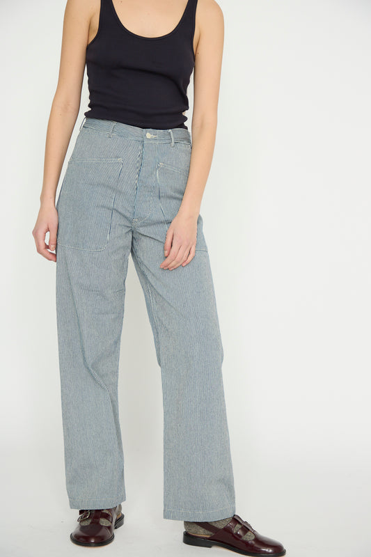 A model in a black tank top and As Ever's Brancusi Pant in Engineer Stripe, featuring a high-waisted, wide-leg silhouette, complemented by brown shoes, stands against a plain white background.