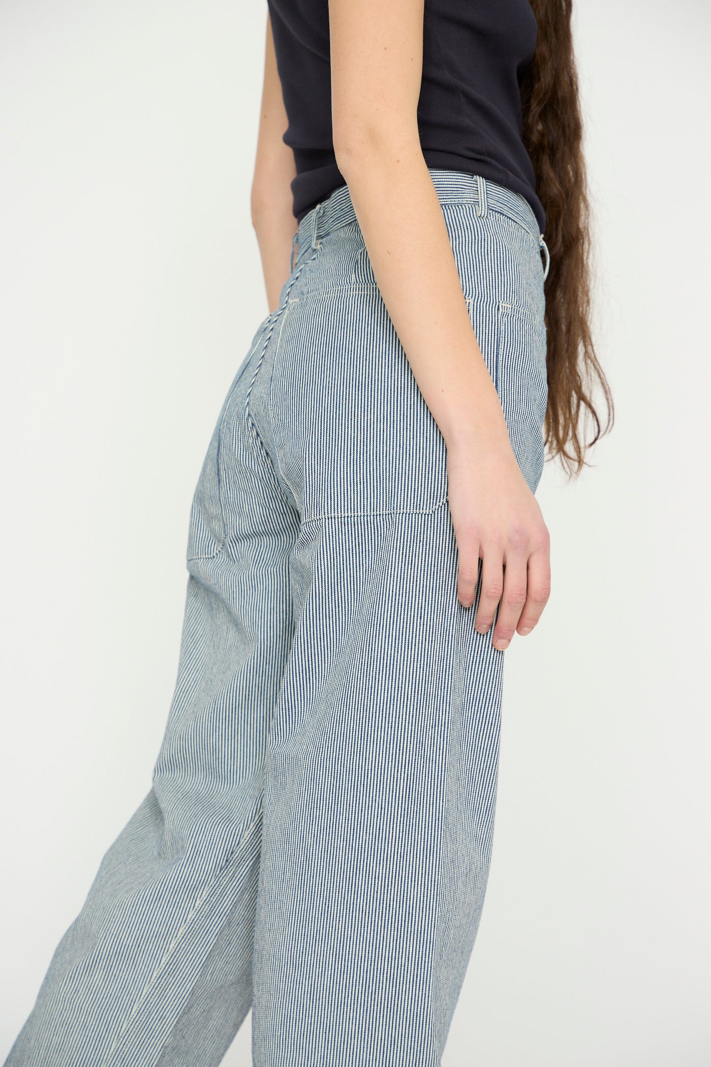 A woman with long hair, wearing a black top and As Ever's Brancusi Pant in Engineer Stripe, is shown from the side against a plain backdrop, highlighting its wide-leg silhouette reminiscent of 1940s vintage style.