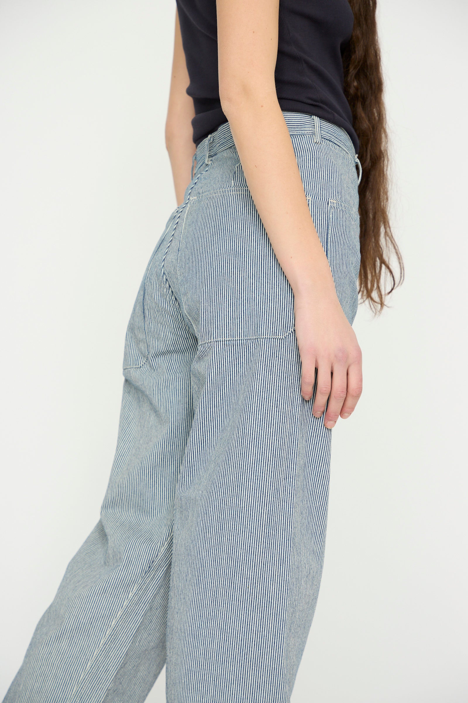 A woman with long hair, wearing a black top and As Ever's Brancusi Pant in Engineer Stripe, is shown from the side against a plain backdrop, highlighting its wide-leg silhouette reminiscent of 1940s vintage style.