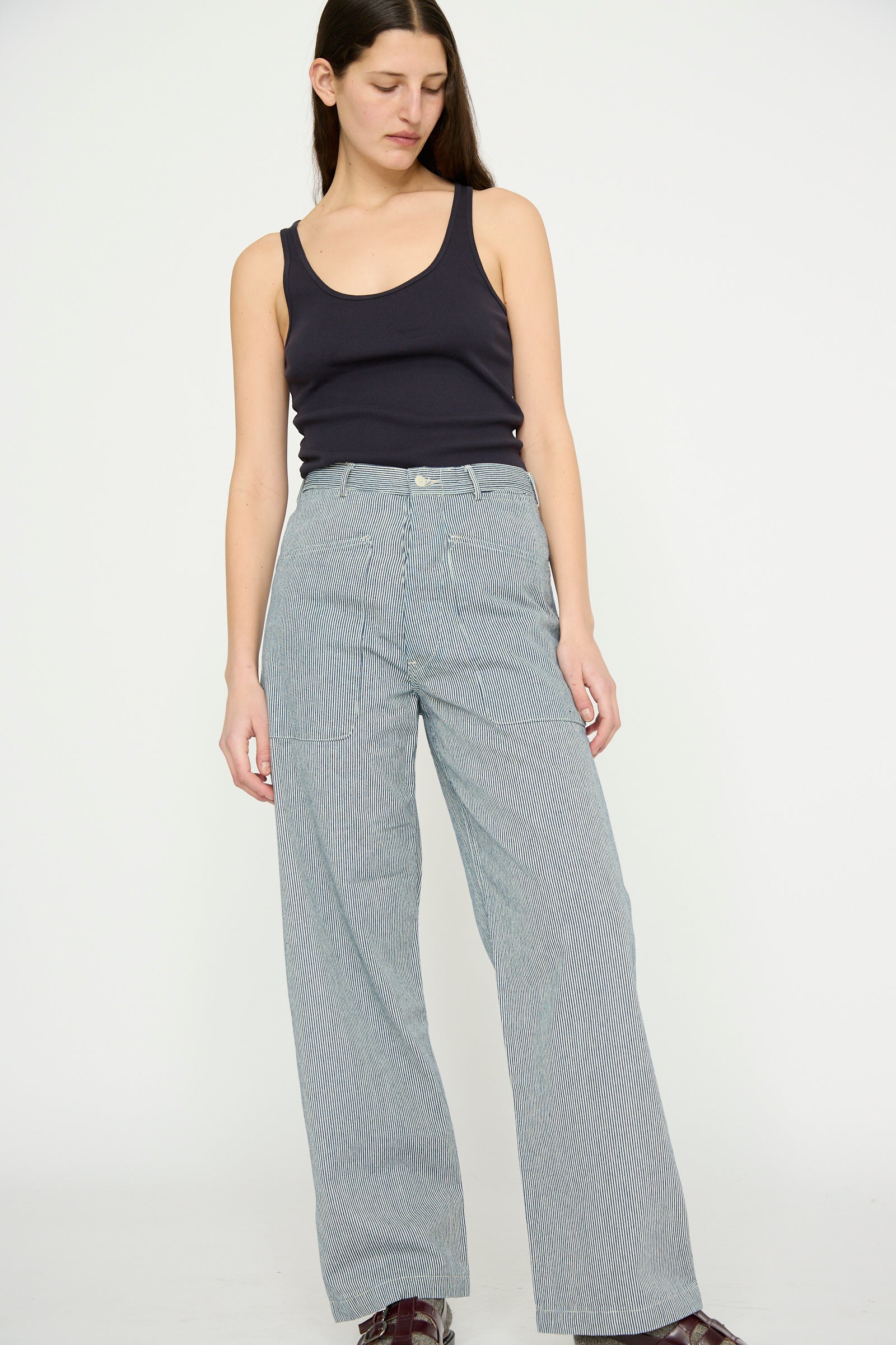 Wearing the As Ever Brancusi Pant in Engineer Stripe with a black tank top, a person exudes a touch of 1940s vintage style against a plain background.