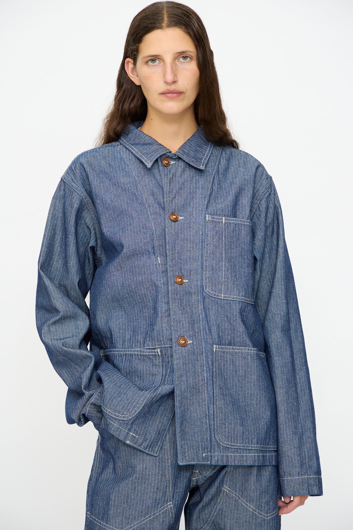 A person wears the As Ever Henri Jacket in HBT Denim, a loose-fitting unisex work jacket with large front pockets and brown buttons, paired with matching denim pants—modern style inspired by 1940s military jackets.