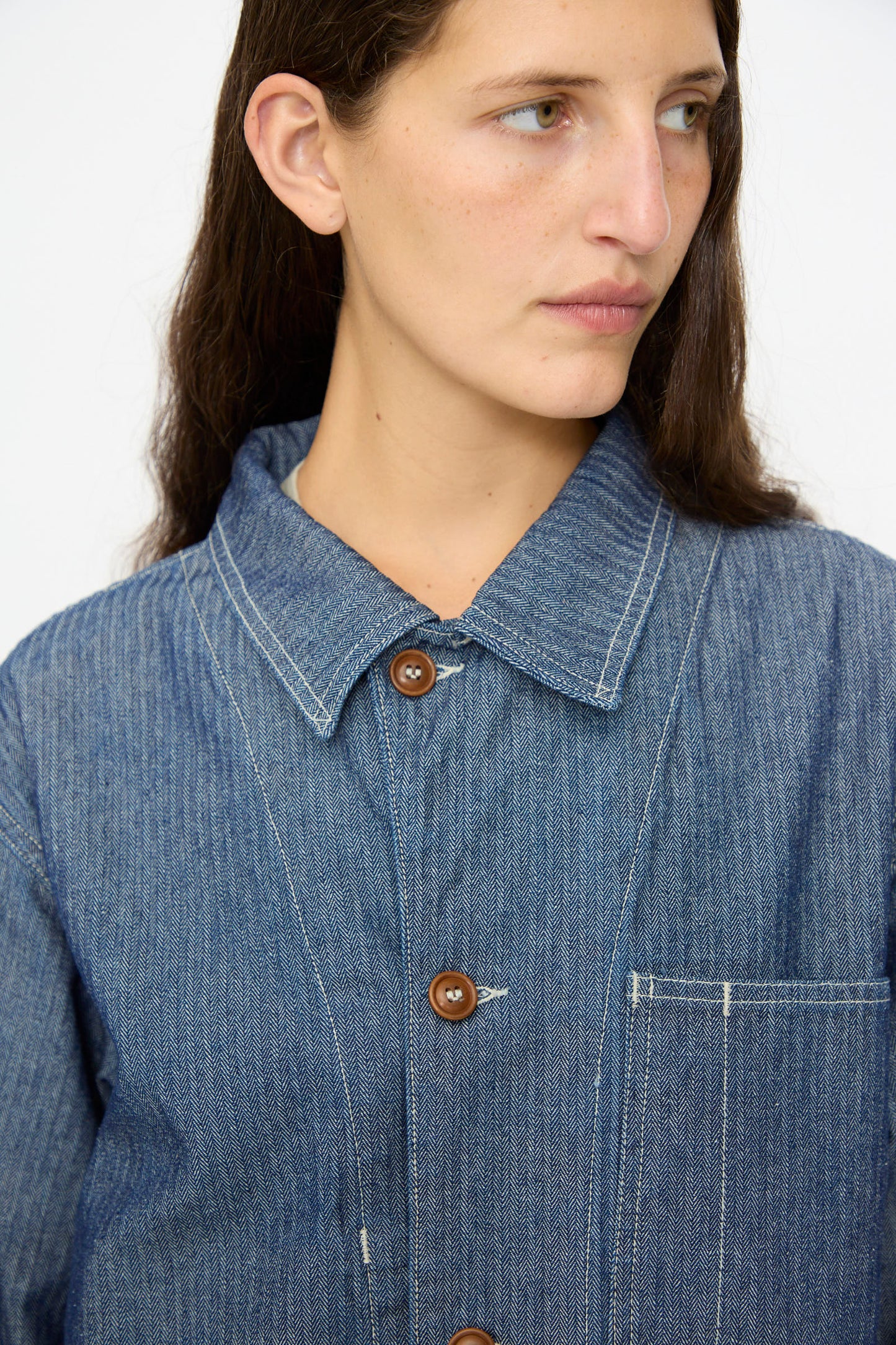 A person wears the As Ever Henri Jacket in HBT Denim—unisex, featuring blue herringbone twill, brown buttons, and a large front pocket—inspired by classic 1940s military jackets.