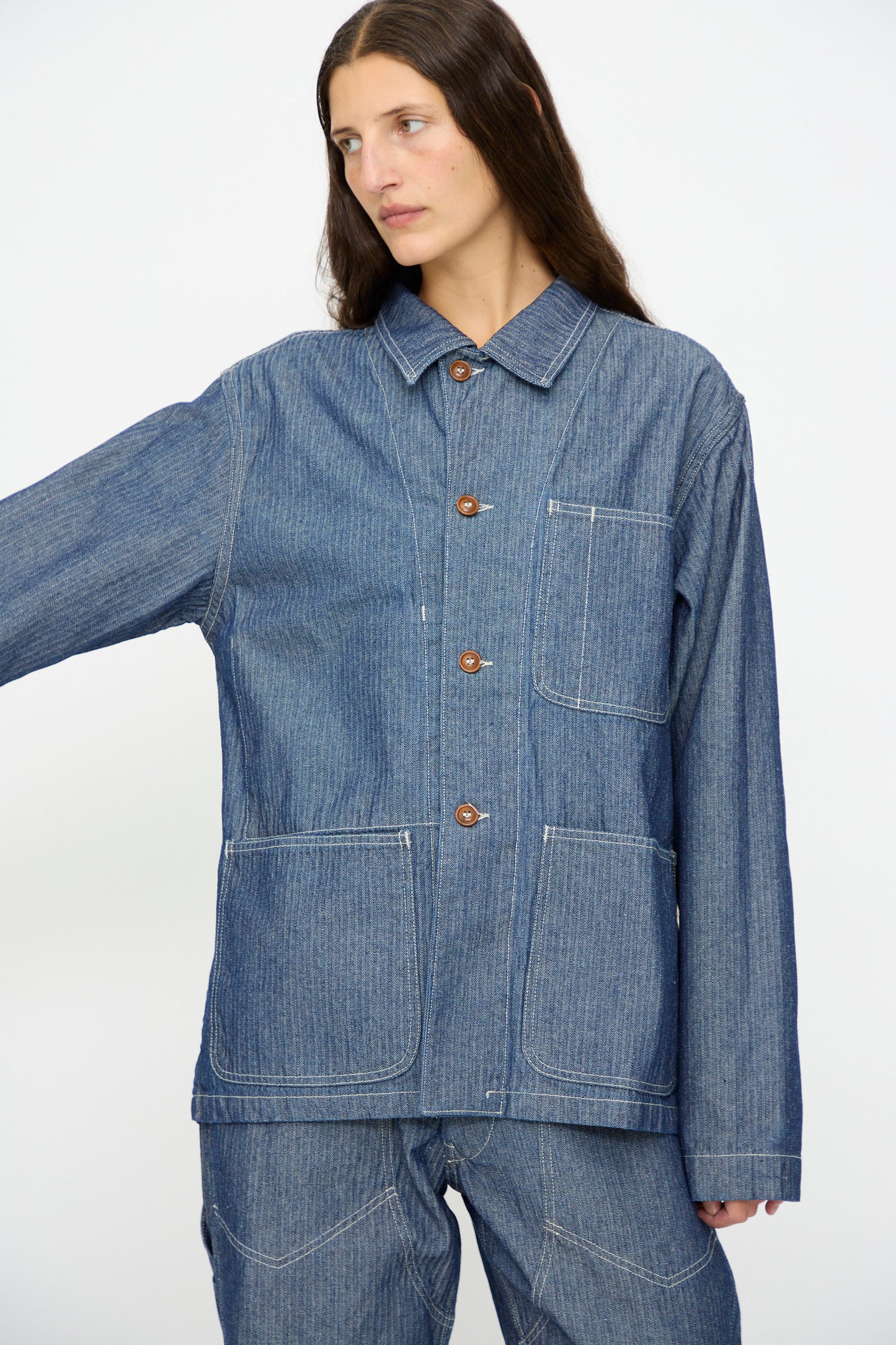 Someone wears the As Ever Henri Jacket in HBT Denim, featuring large front pockets and brown buttons, paired with matching denim pants, standing against a plain background.