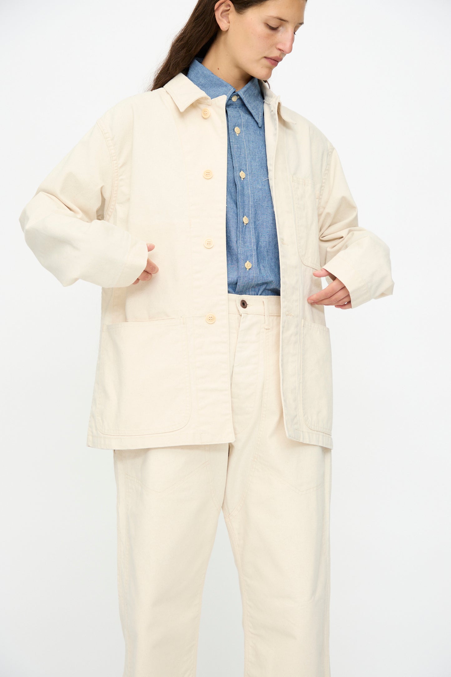 A person wears the As Ever Henri Jacket in Natural over a blue button-up shirt and pants, standing against a plain white background.