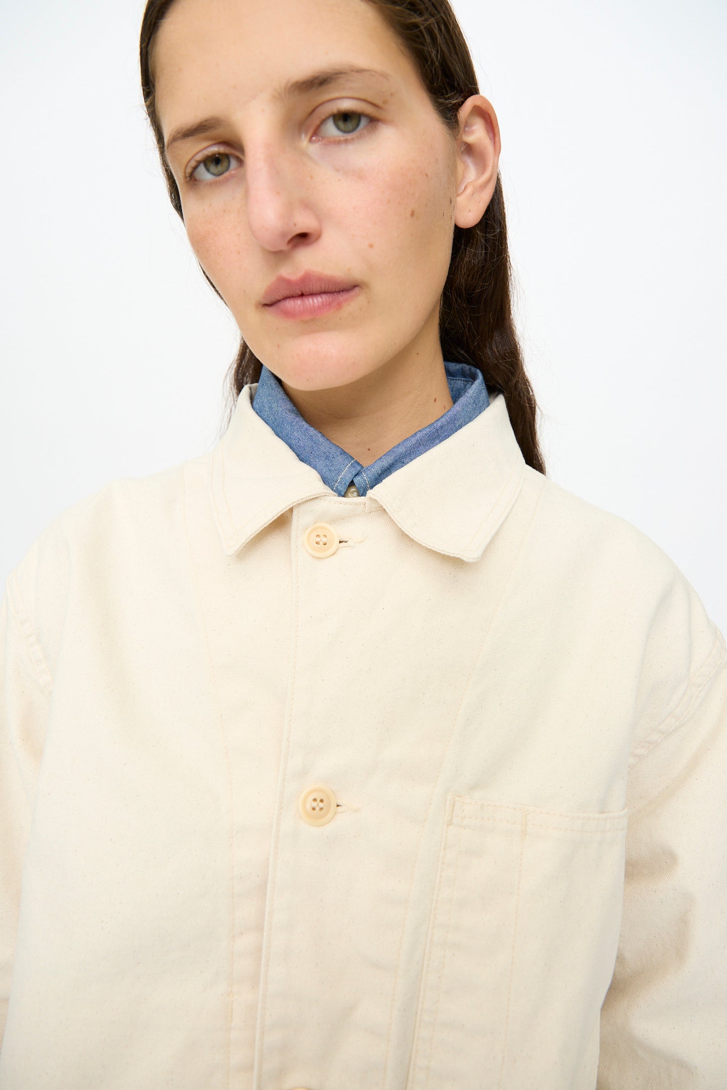 A person with long brown hair wears the As Ever Henri Jacket in Natural over a blue shirt, standing against a plain white background.