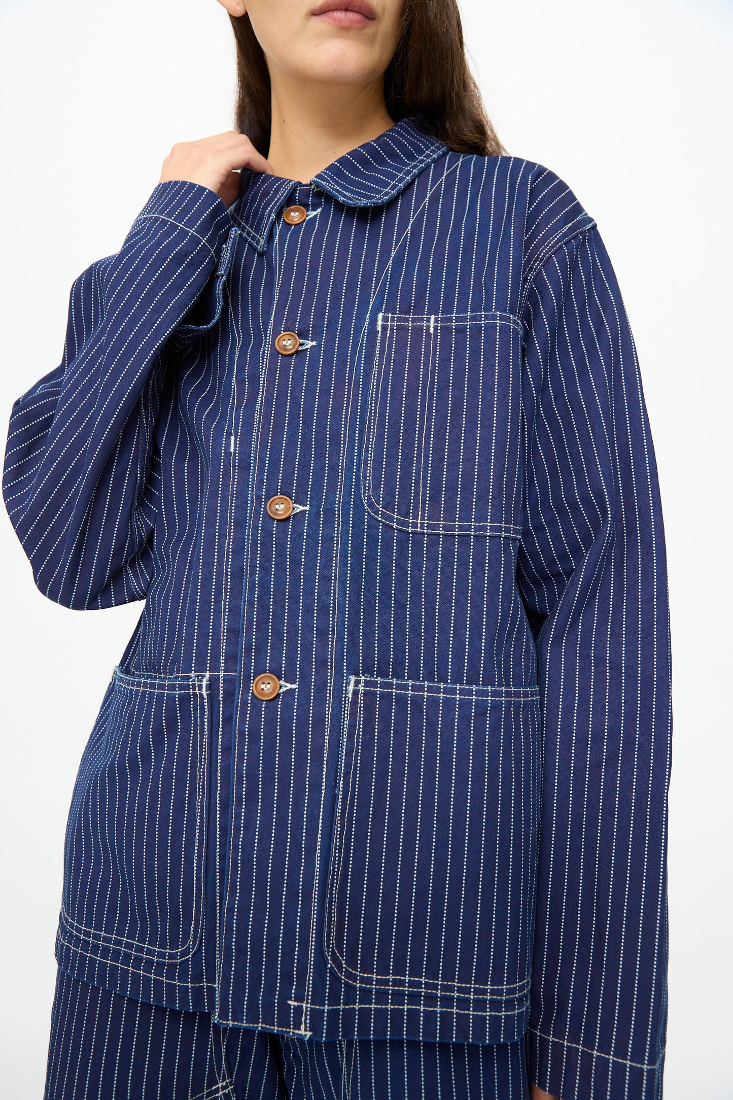 Someone stands against a plain white background wearing the As Ever Henri Jacket in Wabash, featuring indigo fabric with pinstripes and large front pockets.