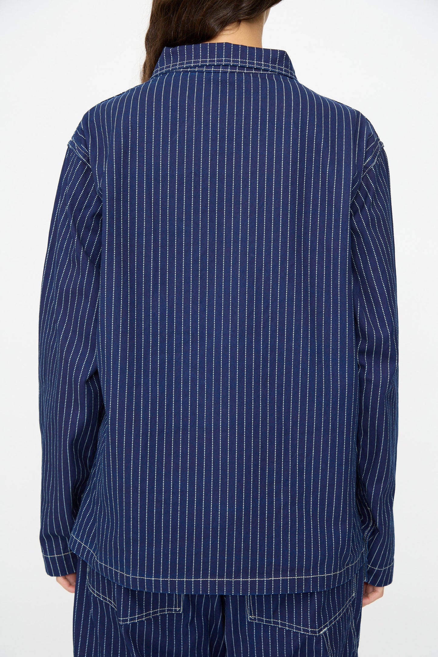 A person is seen from the back wearing the As Ever Henri Jacket in Wabash, featuring indigo fabric with white vertical pinstripes, paired with matching blue pants for a utilitarian look.