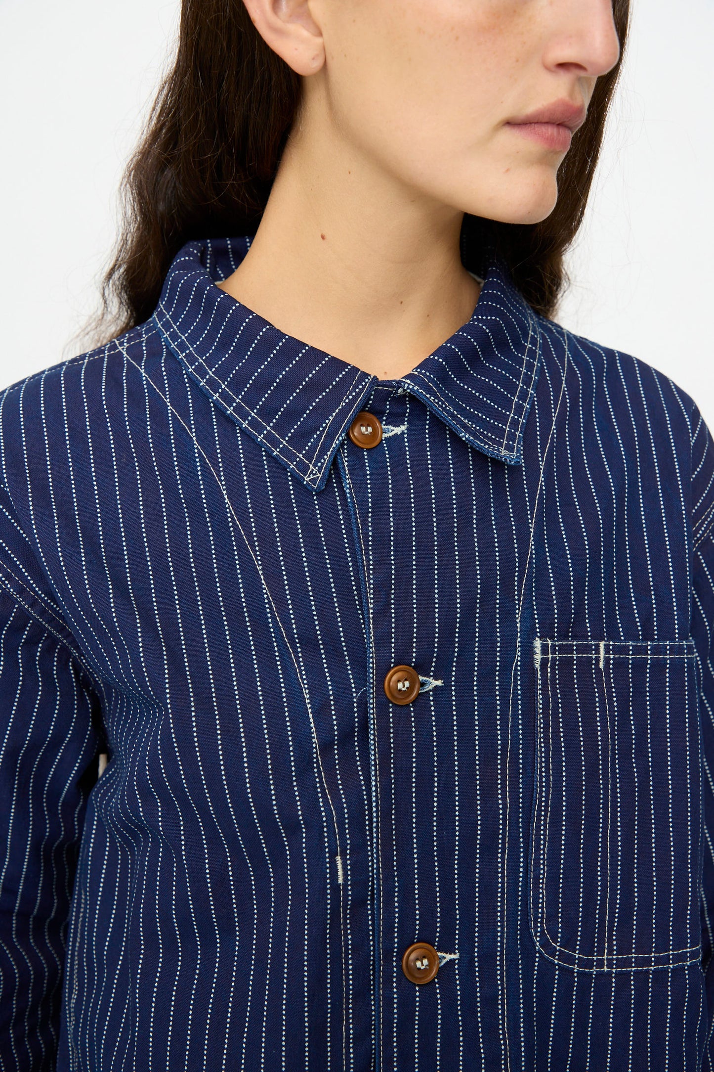 A person shown from the shoulders up and facing sideways wears the As Ever Henri Jacket in Wabash, featuring blue and white pinstripes, a collar, chest pocket, and classic workwear-inspired style.