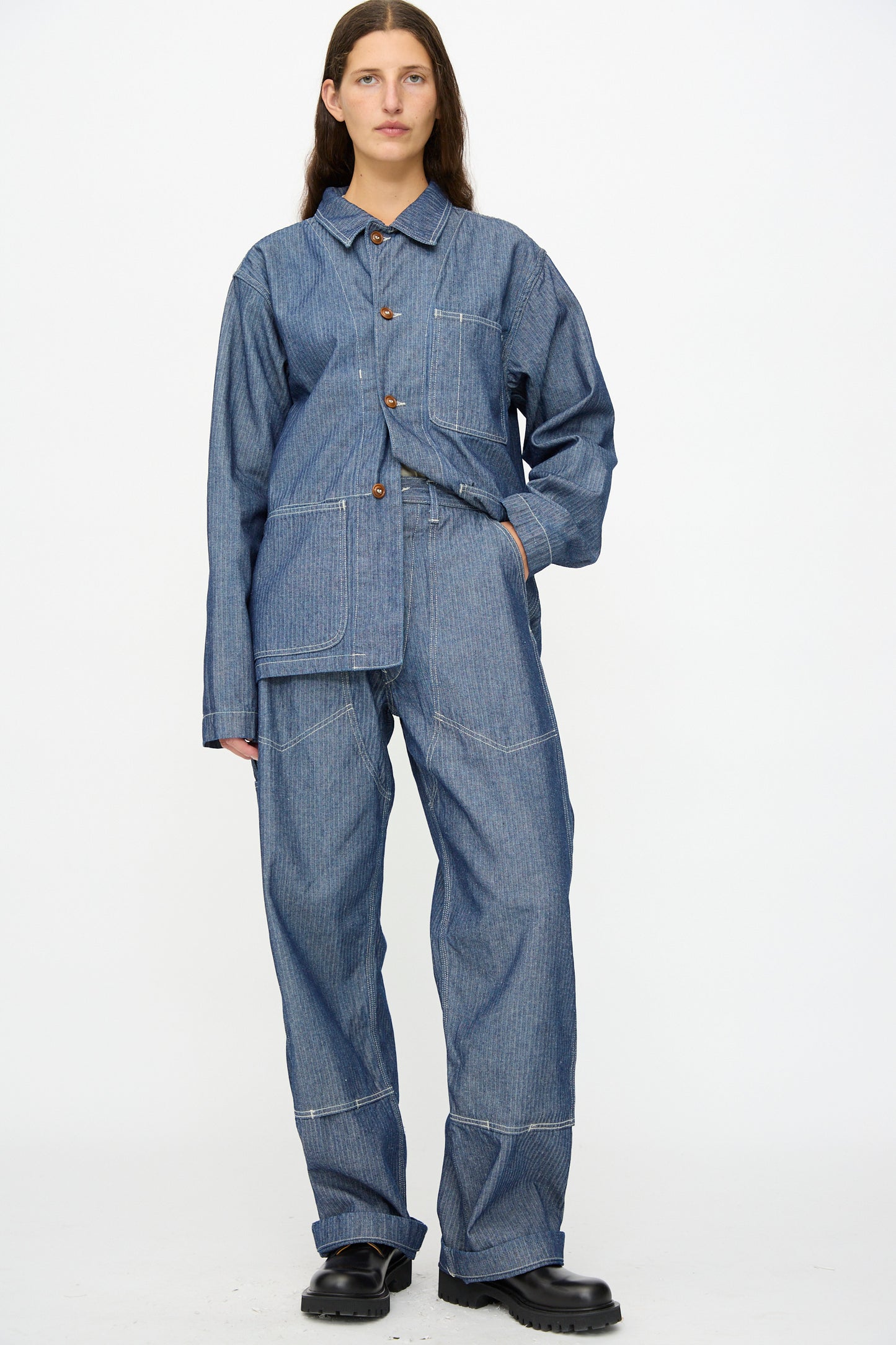 A person stands against a plain background wearing the As Ever Henri Pant in HBT Denim with a matching blue denim workwear-style jacket and black shoes.