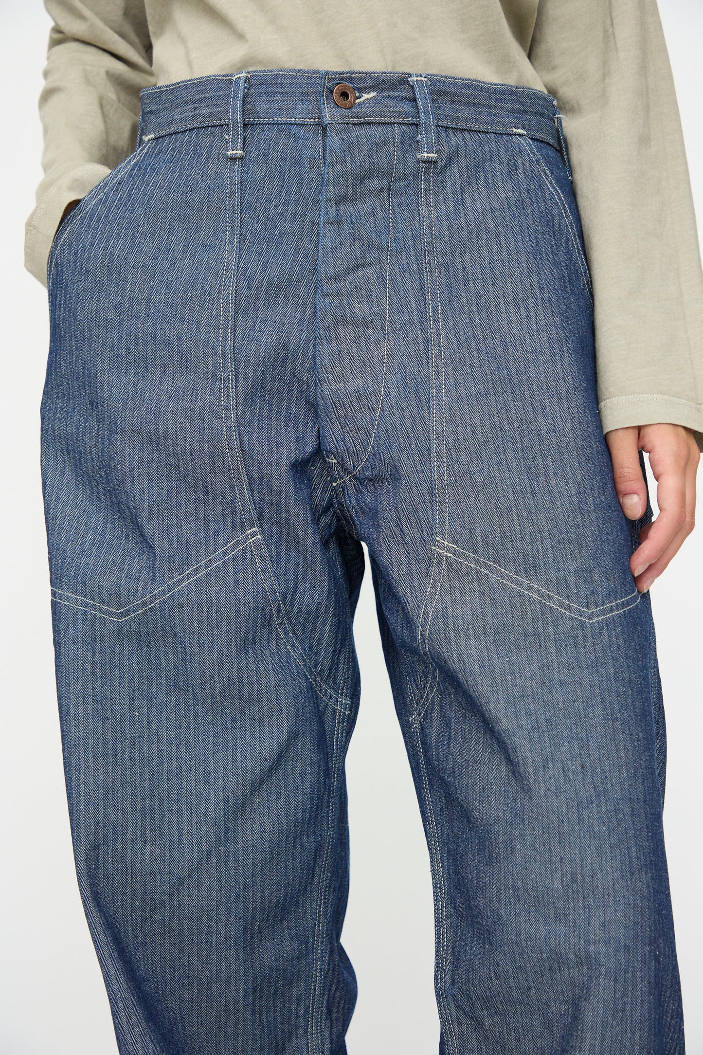 A person wears a light long-sleeve shirt with the Henri Pant in HBT Denim by As Ever—high-waisted, vintage military pants made from blue denim with visible white stitching.