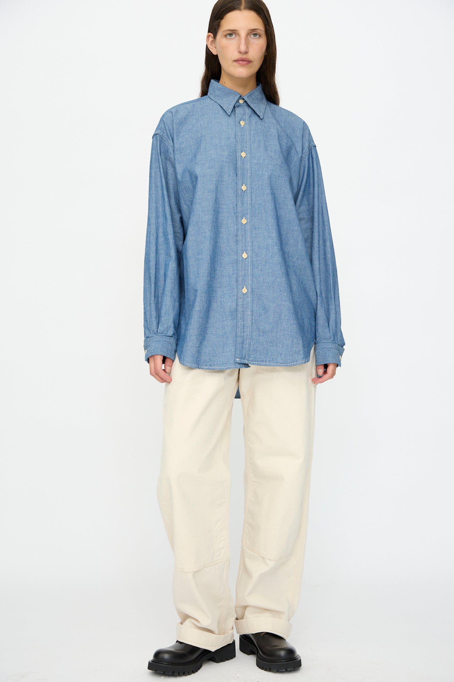 Person stands against a plain background wearing a loose blue denim button-up shirt, As Ever Henri Pant in Natural (off-white), and black shoes.