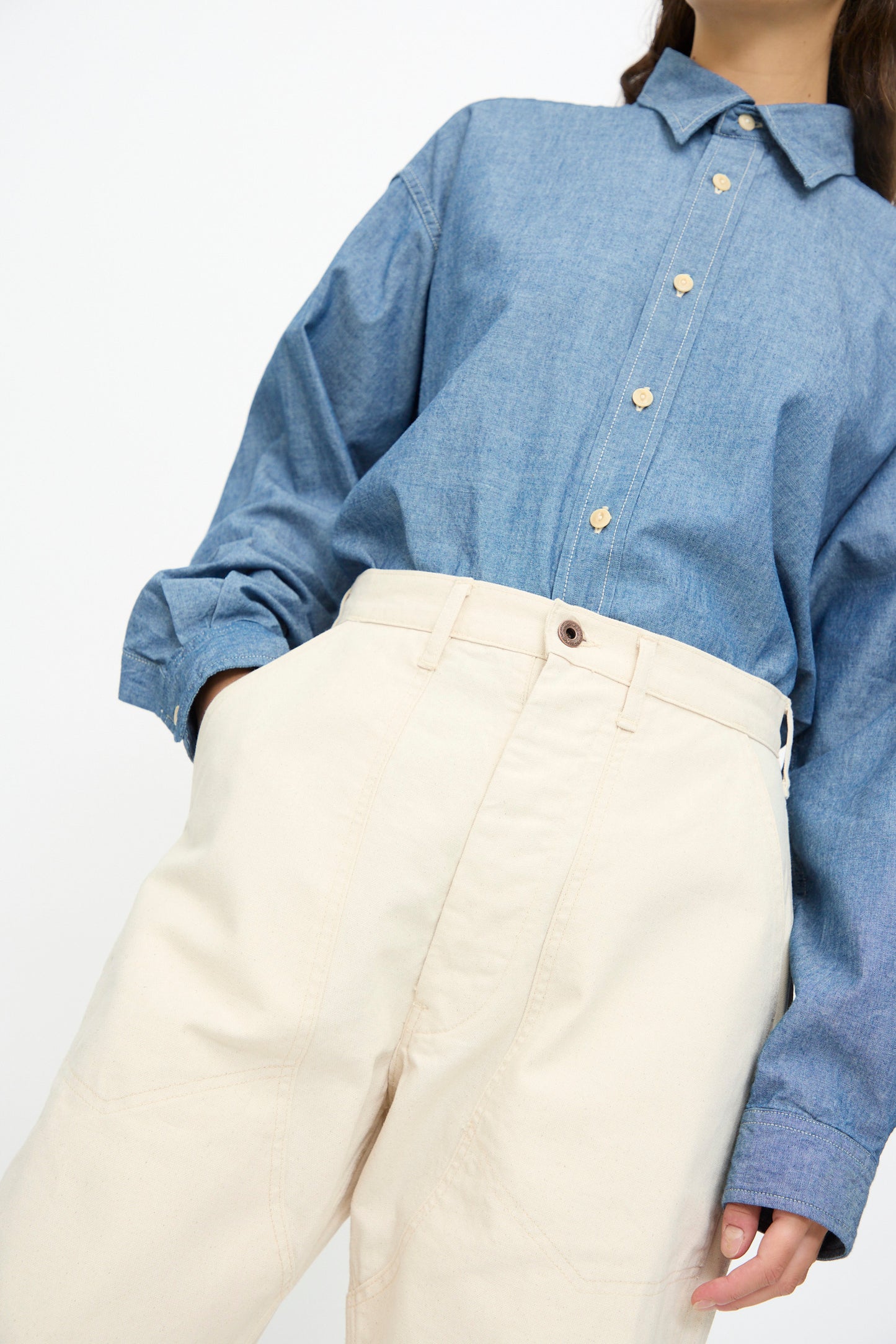 A person wears the As Ever Henri Pant in Natural—high-waisted and oversized—paired with a blue button-up shirt, tucked in with one hand in their pocket. The face is partially out of frame.